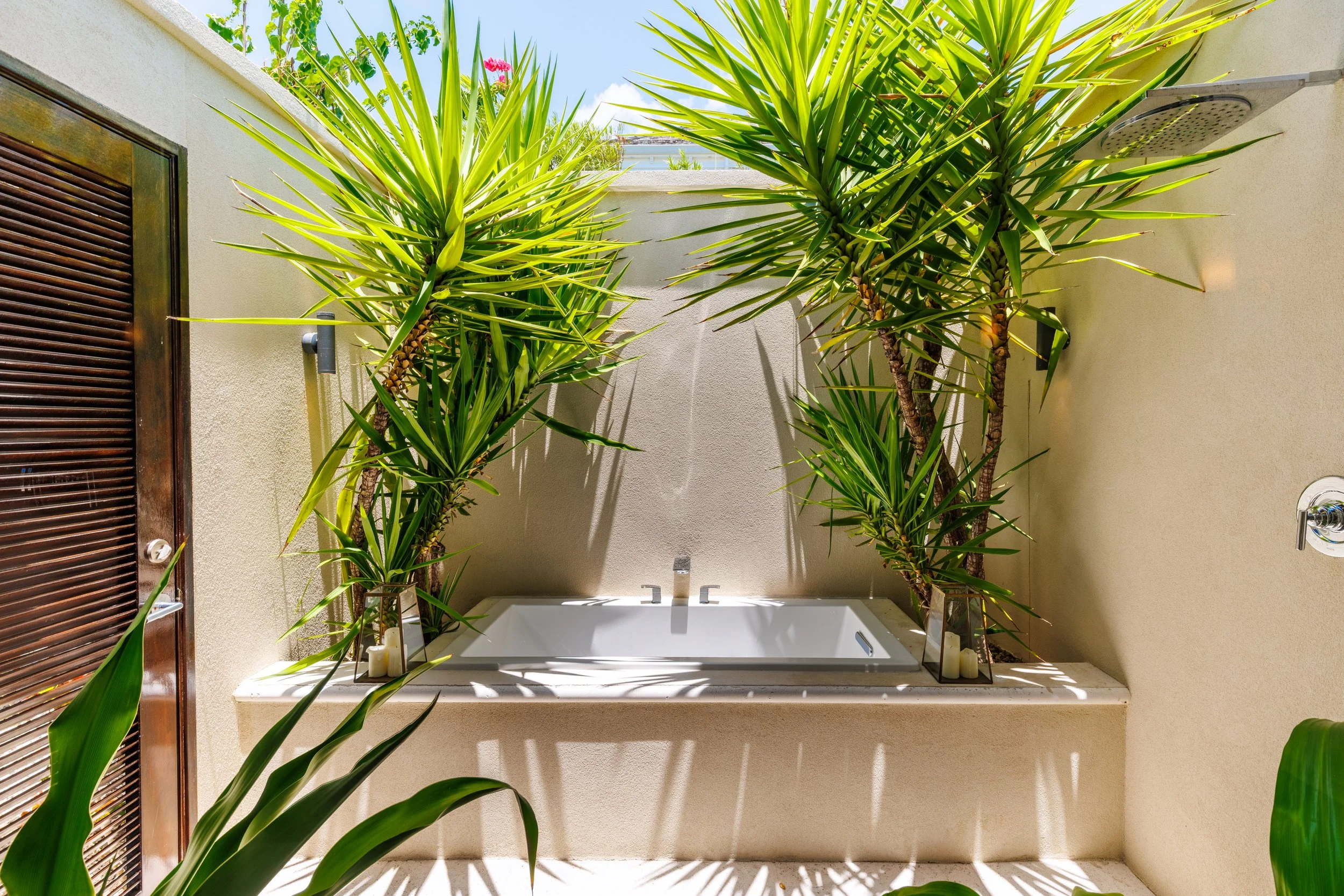 An outdoor shower area with a bathtub and tall green plants on either side, set against beige walls and a bright sky.