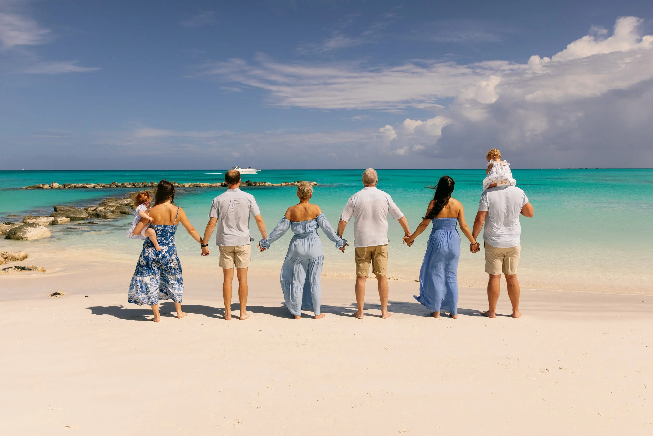 A group of eight people holding hands and walking along the beach near the ocean, with some holding children, under a partly cloudy sky, with a boat in the distance.