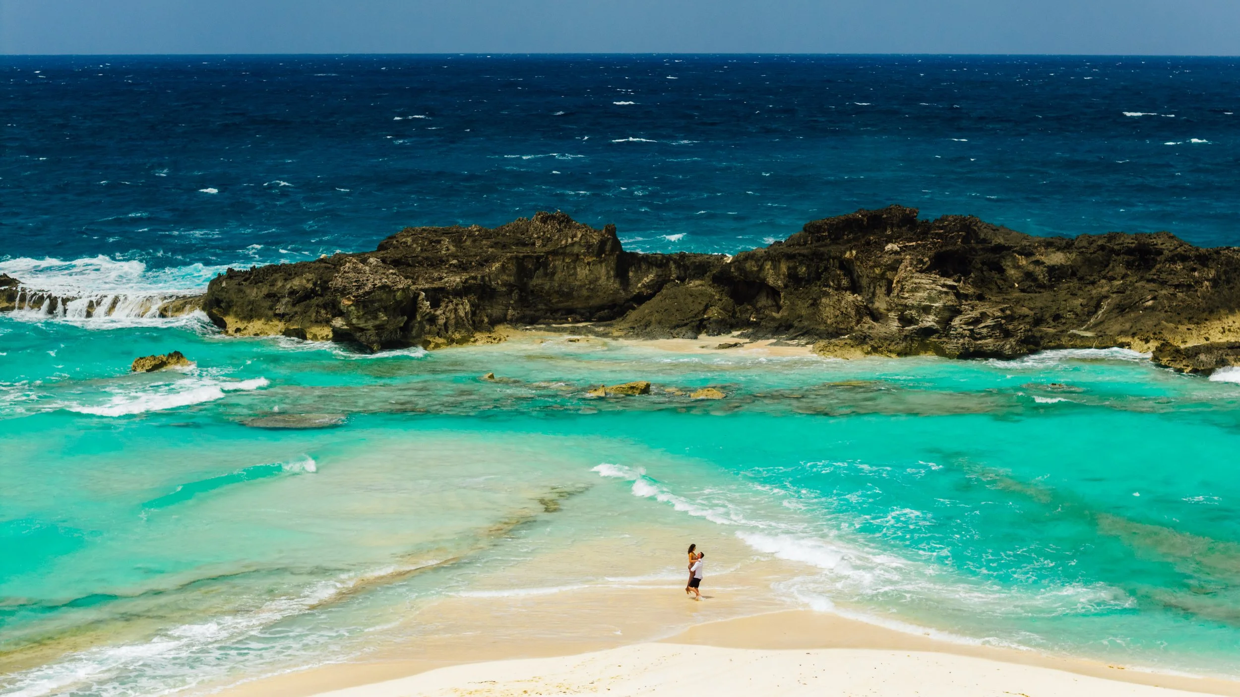 A couple embracing on a sandy beach with turquoise water and rocky cliffs in the background under a clear sky.
