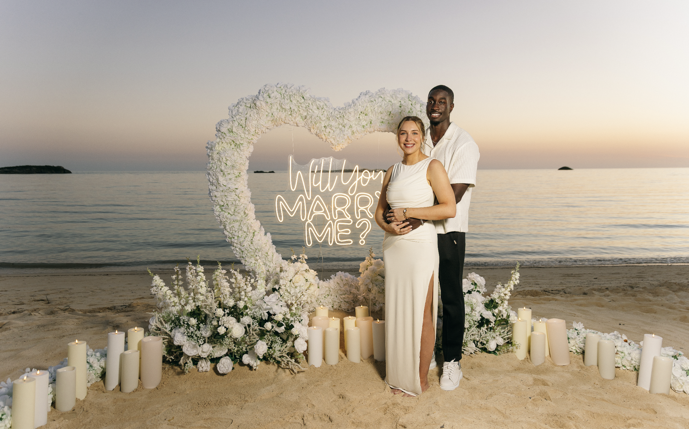 A couple stands on a beach at sunset, smiling, in front of a flower heart arch with a neon sign reading "Will You Marry Me?" surrounded by candles and flowers.