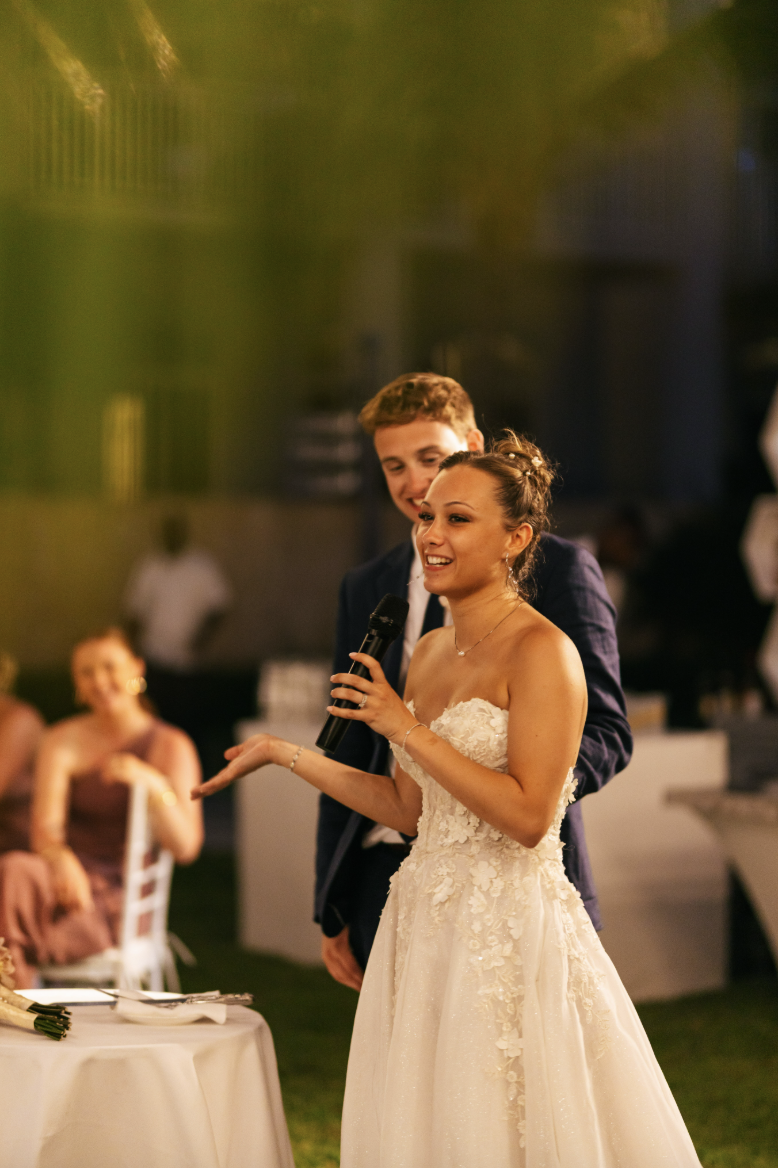 A bride in a white wedding dress with lace details and a groom in a dark suit holding a microphone and giving a speech at a wedding reception. Guests are seated at tables in the background.