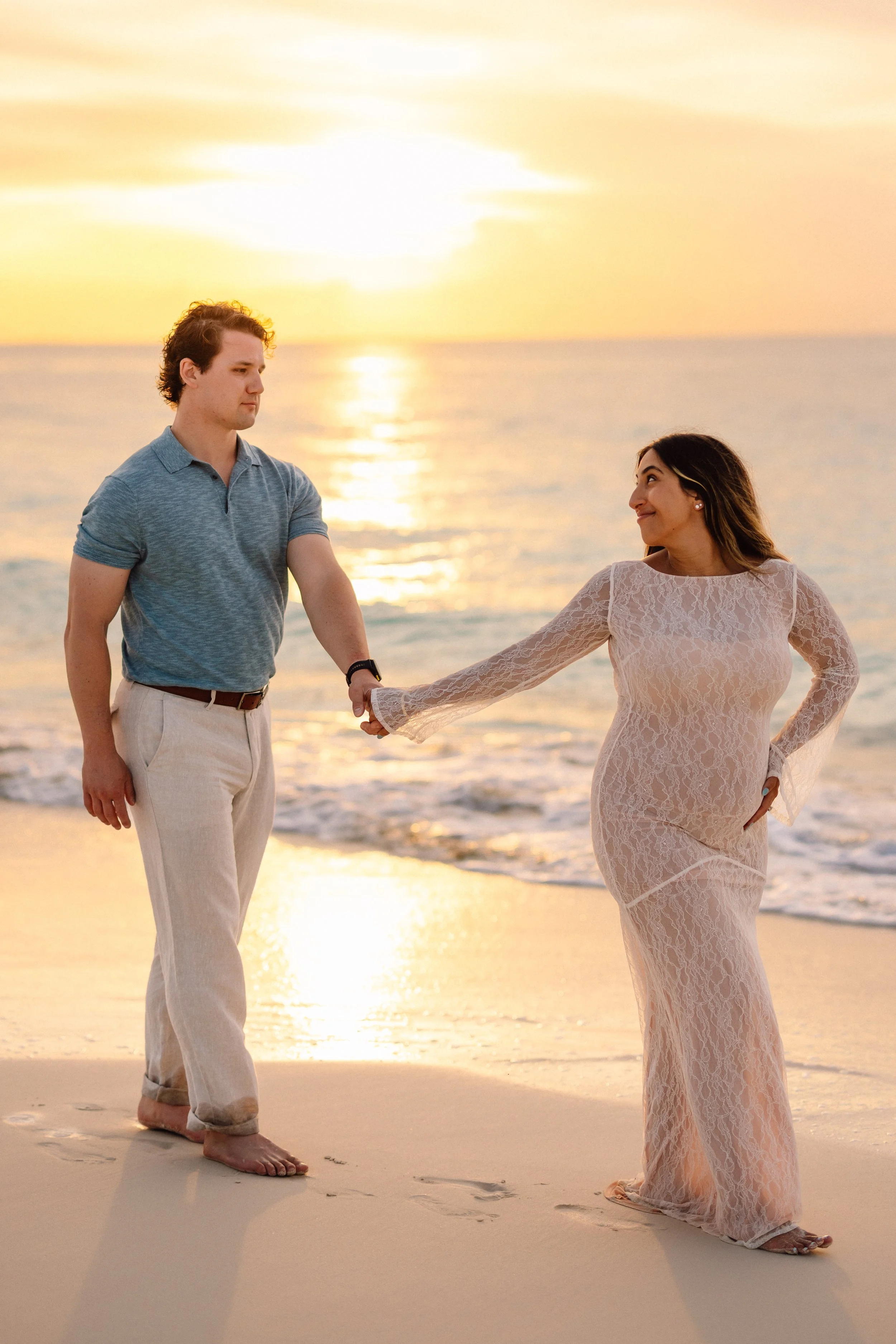 A couple holding hands on the beach at sunset, with the man in a blue polo shirt and beige pants, and the woman in a long, sheer lace dress, both barefoot, standing on the sand with the ocean and sunset in the background.