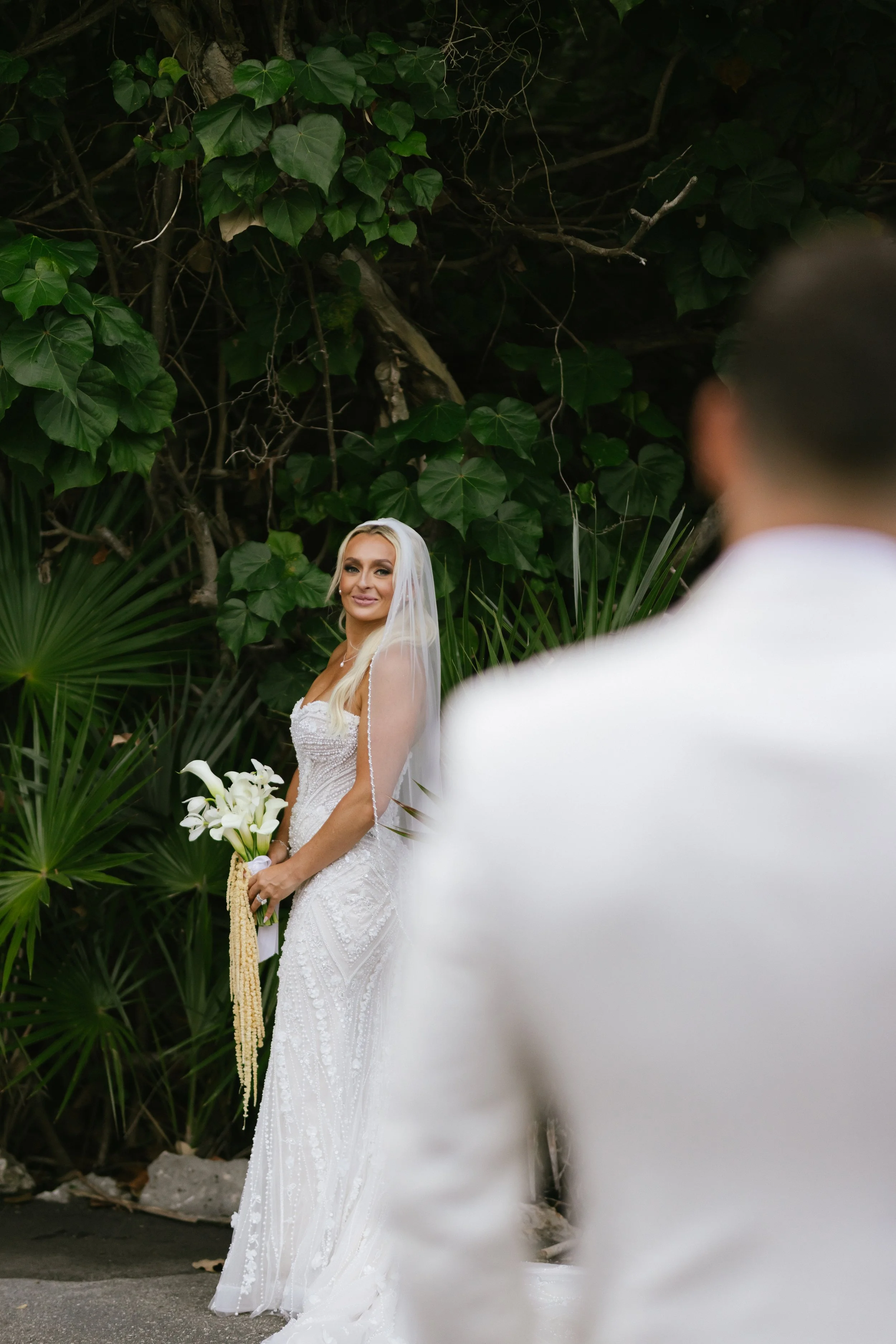 A bride in a white wedding dress and veil holding a bouquet of white flowers, standing outdoors with lush green foliage in the background. A groom in a white suit is partially visible in the foreground.