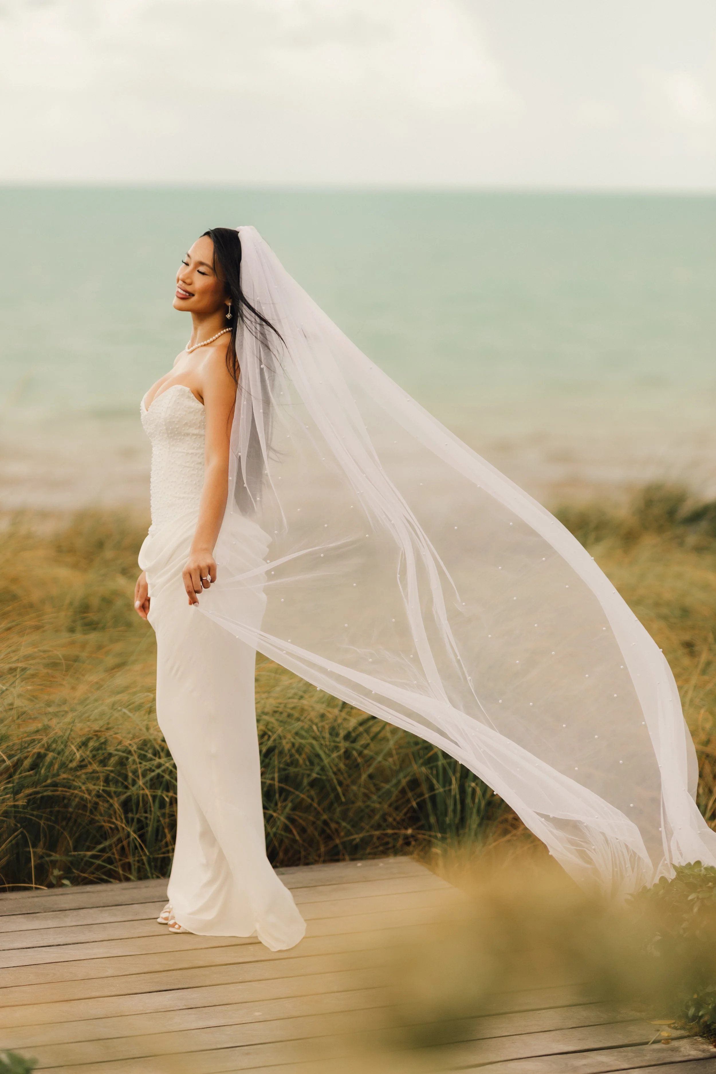 A woman in a white wedding dress and veil standing on a wooden deck near a grassy area with a body of water in the background, smiling and with eyes closed.