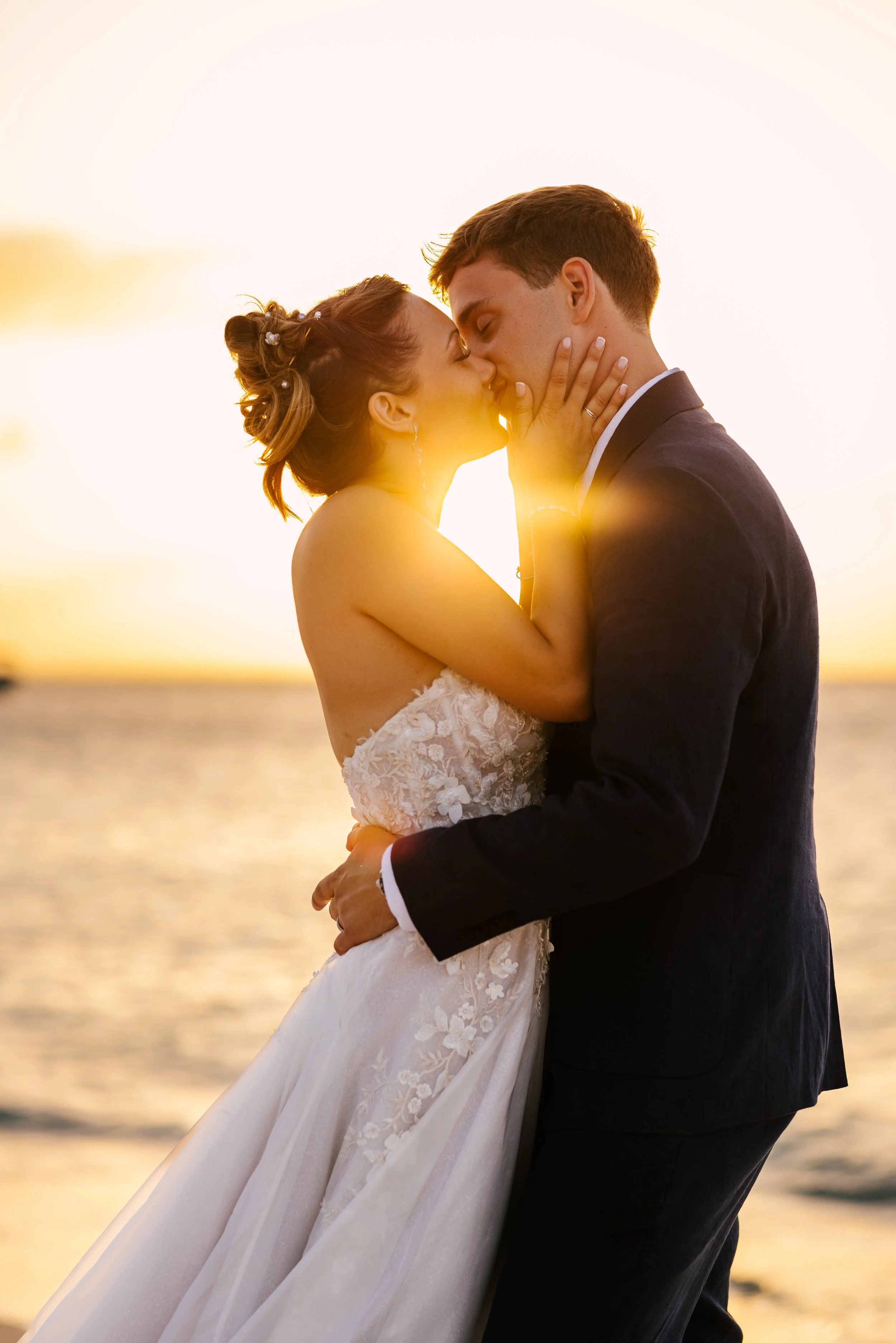 A bride and groom kissing during their wedding at sunset by the water, with the bride wearing a lace gown and the groom in a dark suit.