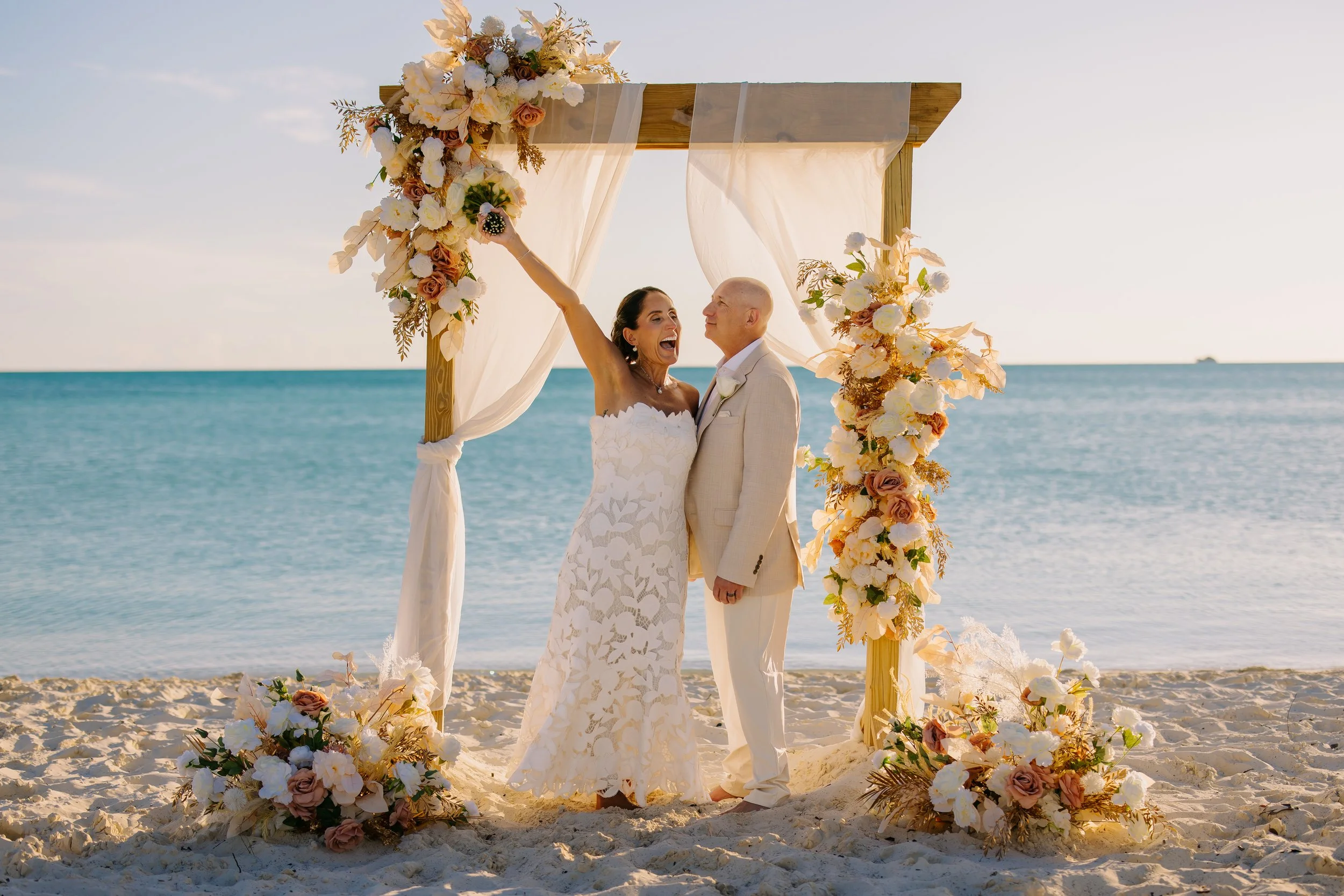 A bride and groom stand under a decorated wedding arch on a beach, with ocean and sky in the background. The arch is adorned with white, peach, and blush flowers and sheer fabric. The bride is wearing a white lace dress and the groom is in a beige su
