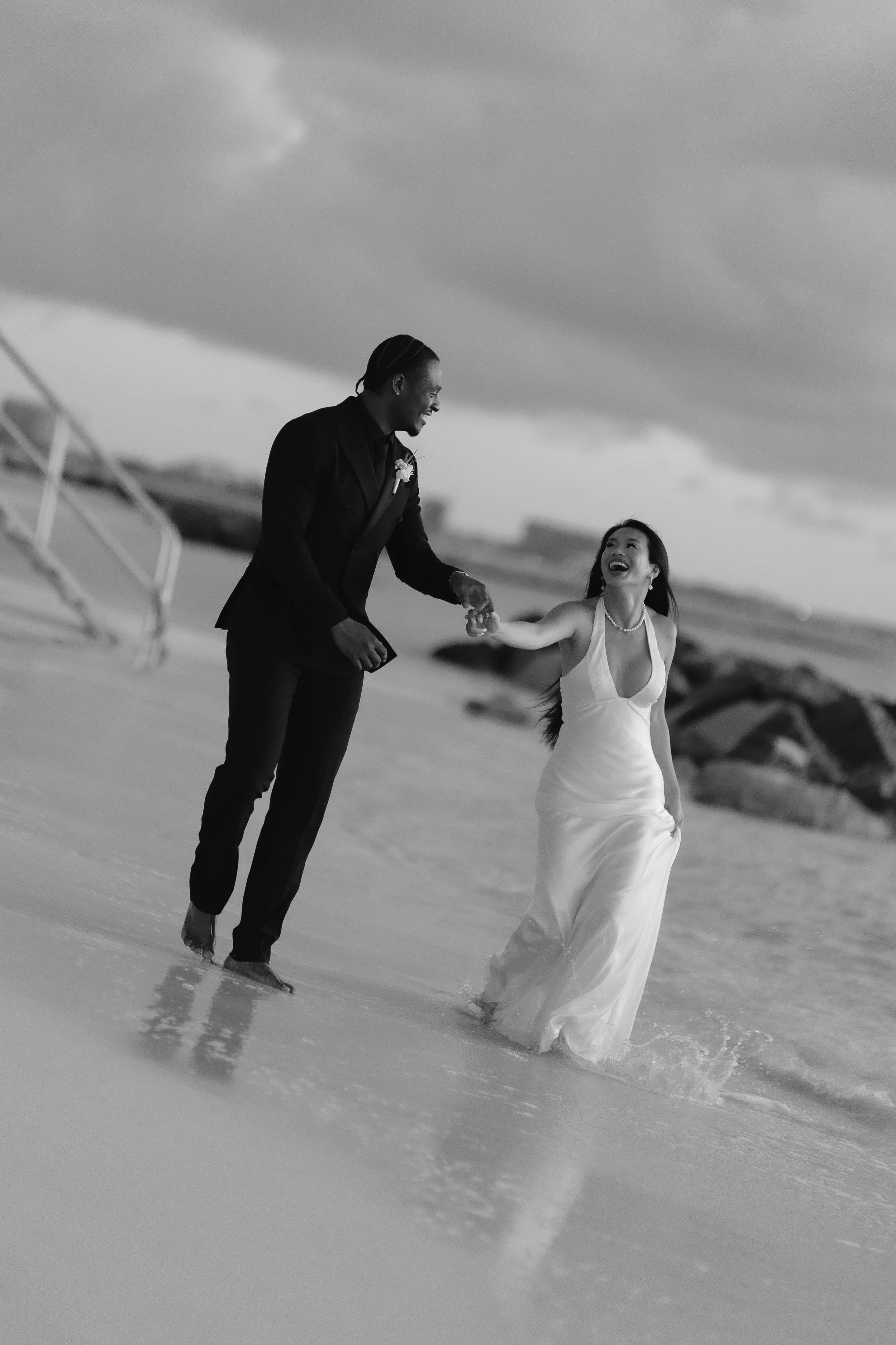 A black and white photo of a couple in wedding attire holding hands and laughing while walking on the beach.