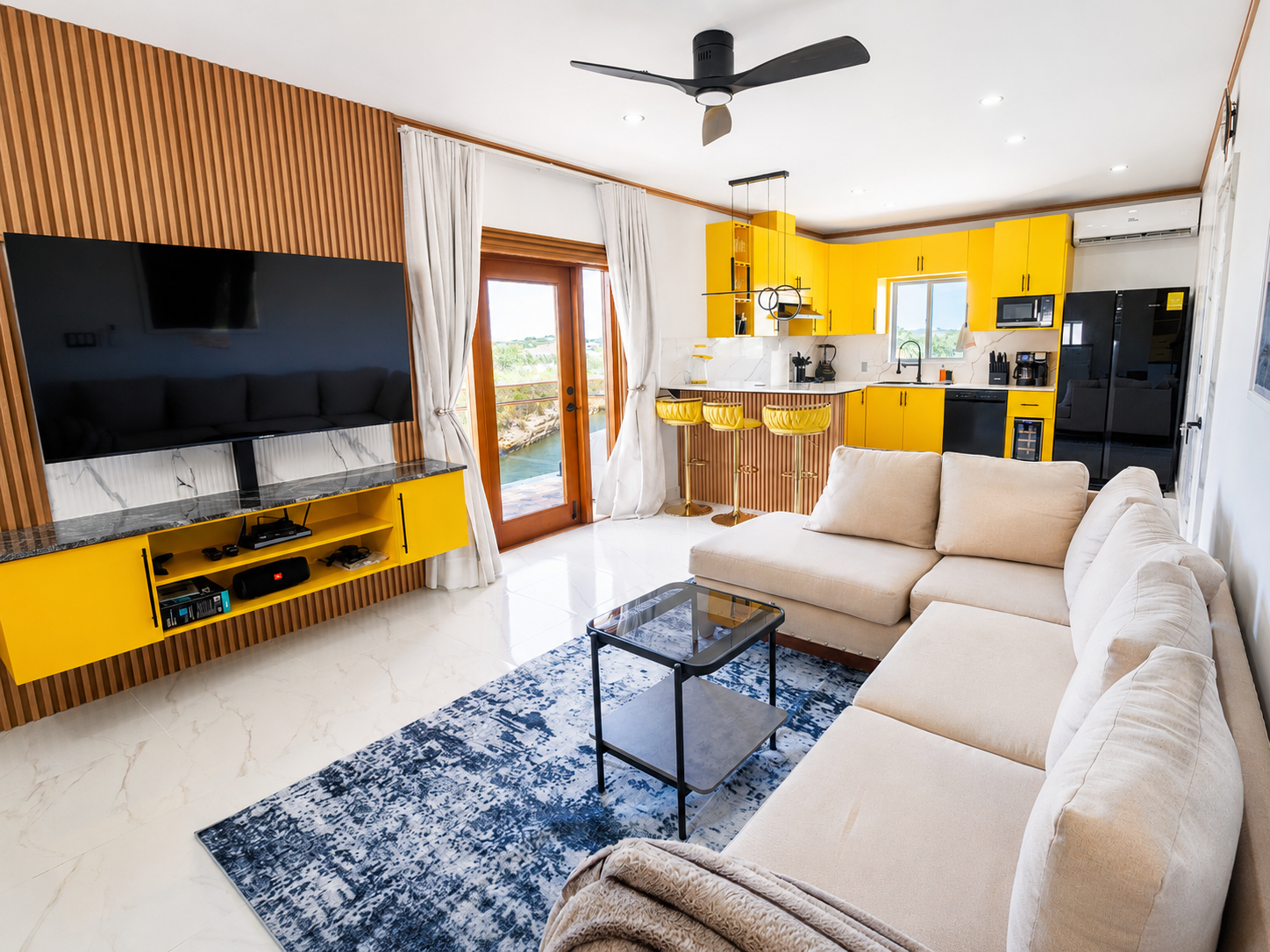 Living room with beige sectional sofa, black coffee table, flat-screen TV on a wooden accent wall, yellow kitchen cabinets, bar stools, and sliding glass door with a water view