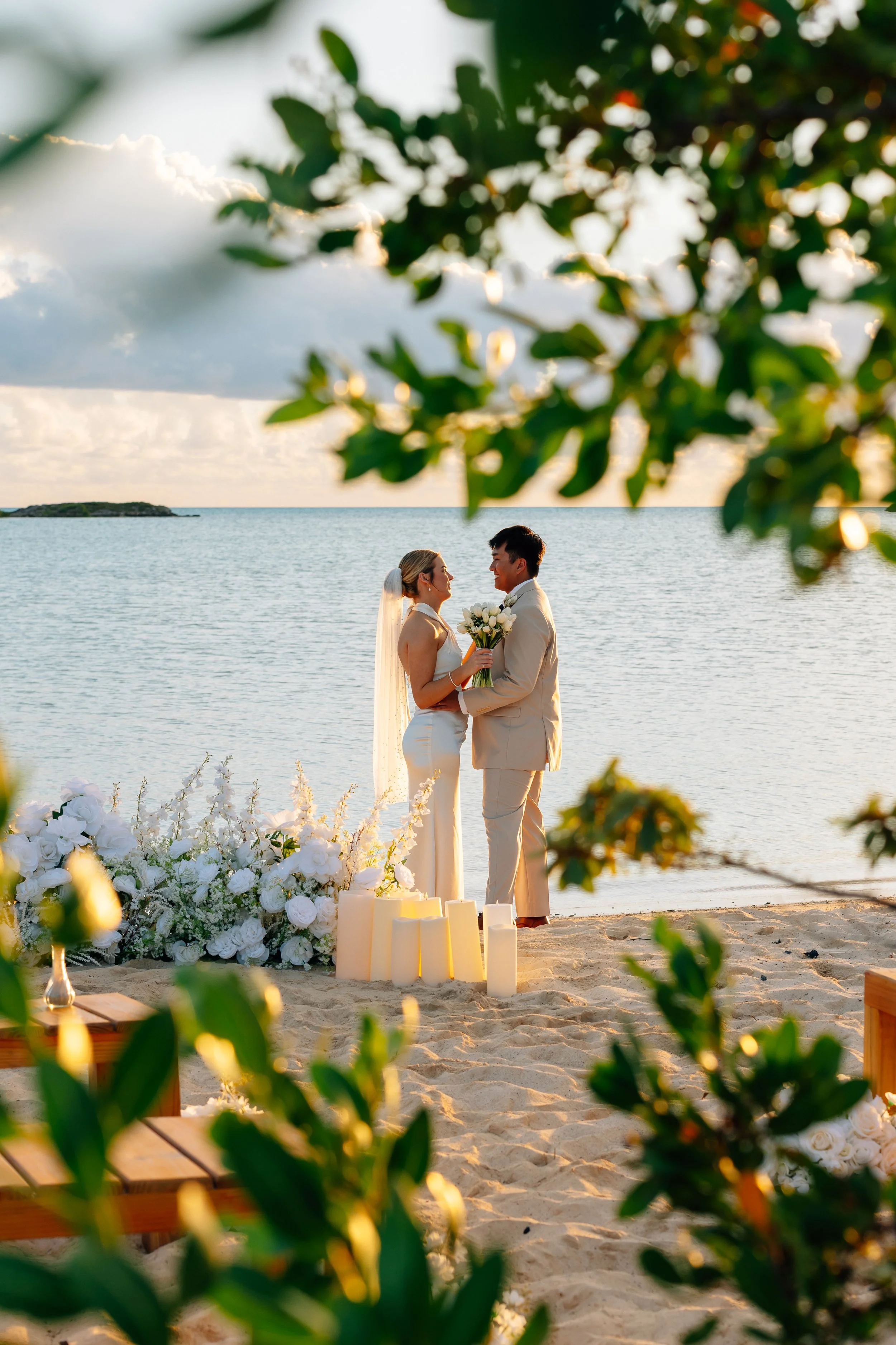 A bride and groom holding hands and holding a bouquet of white flowers during a beach wedding at sunset, viewed through green leaves and white flower arrangements.