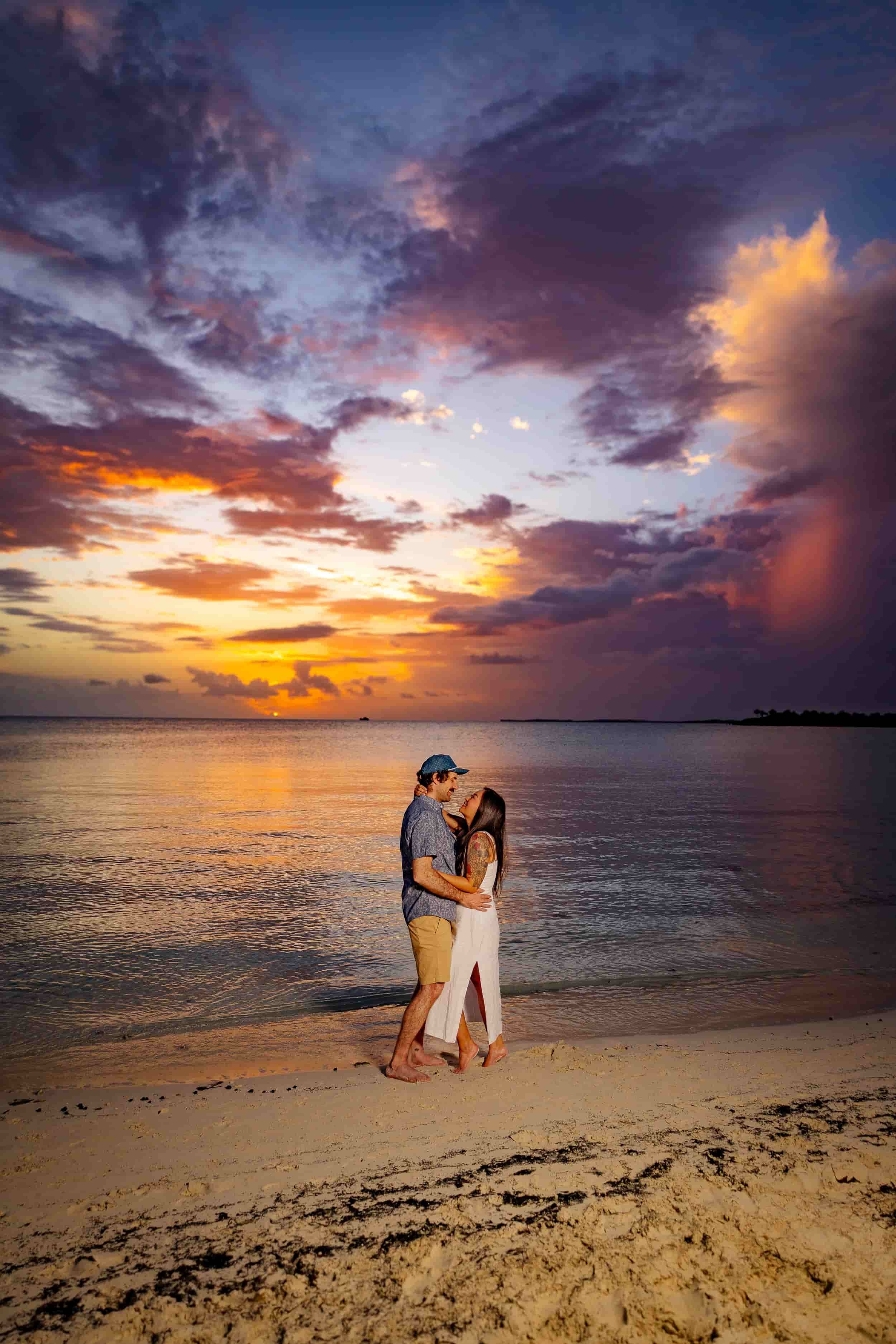 A man and woman embracing on a beach during sunset, with dramatic colorful clouds in the sky and calm water reflecting the sky's colors.