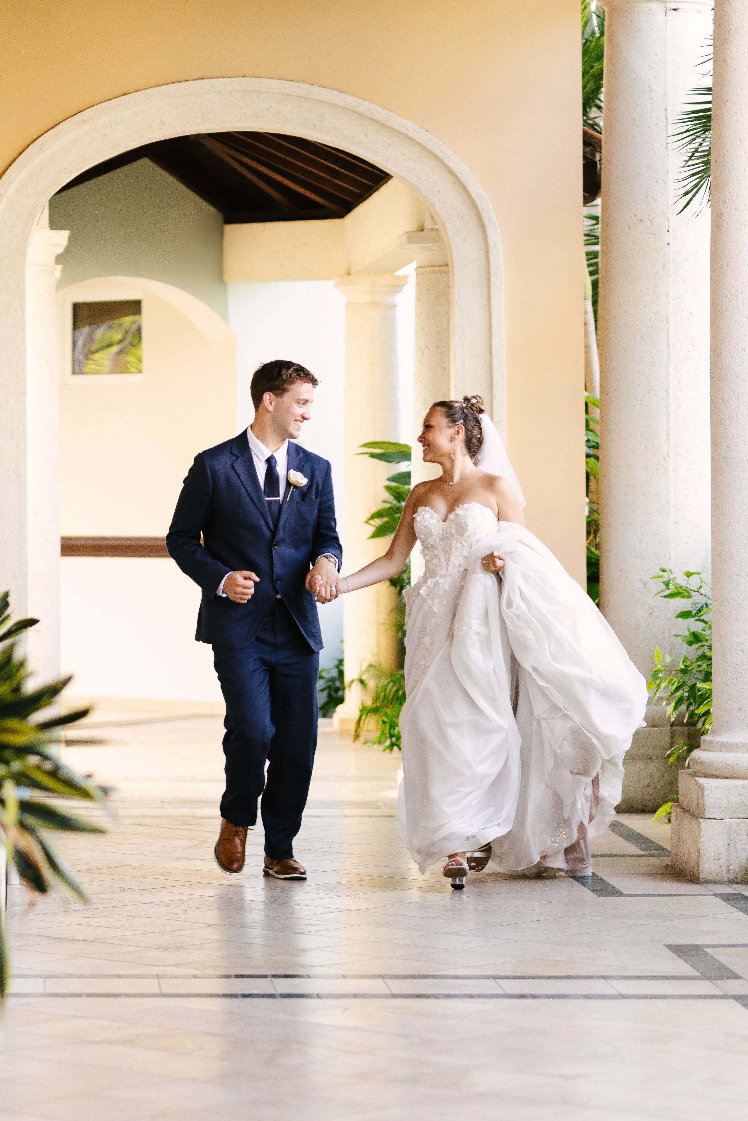 A bride and groom walking hand in hand through a corridor with arches, smiling at each other after their wedding ceremony.