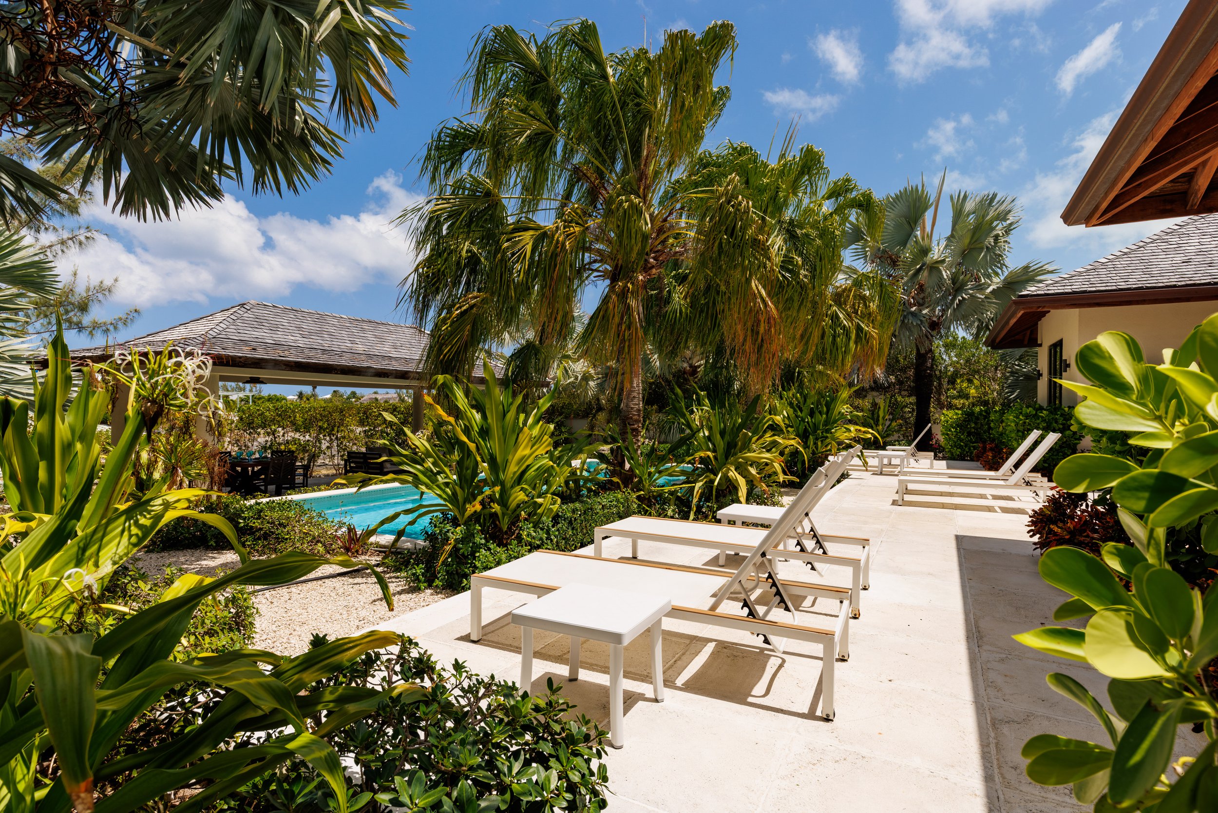 Poolside outdoor area with white lounge chairs, lush green tropical plants, tall palm trees, a thatched gazebo, and a building with a tiled roof under a blue sky.