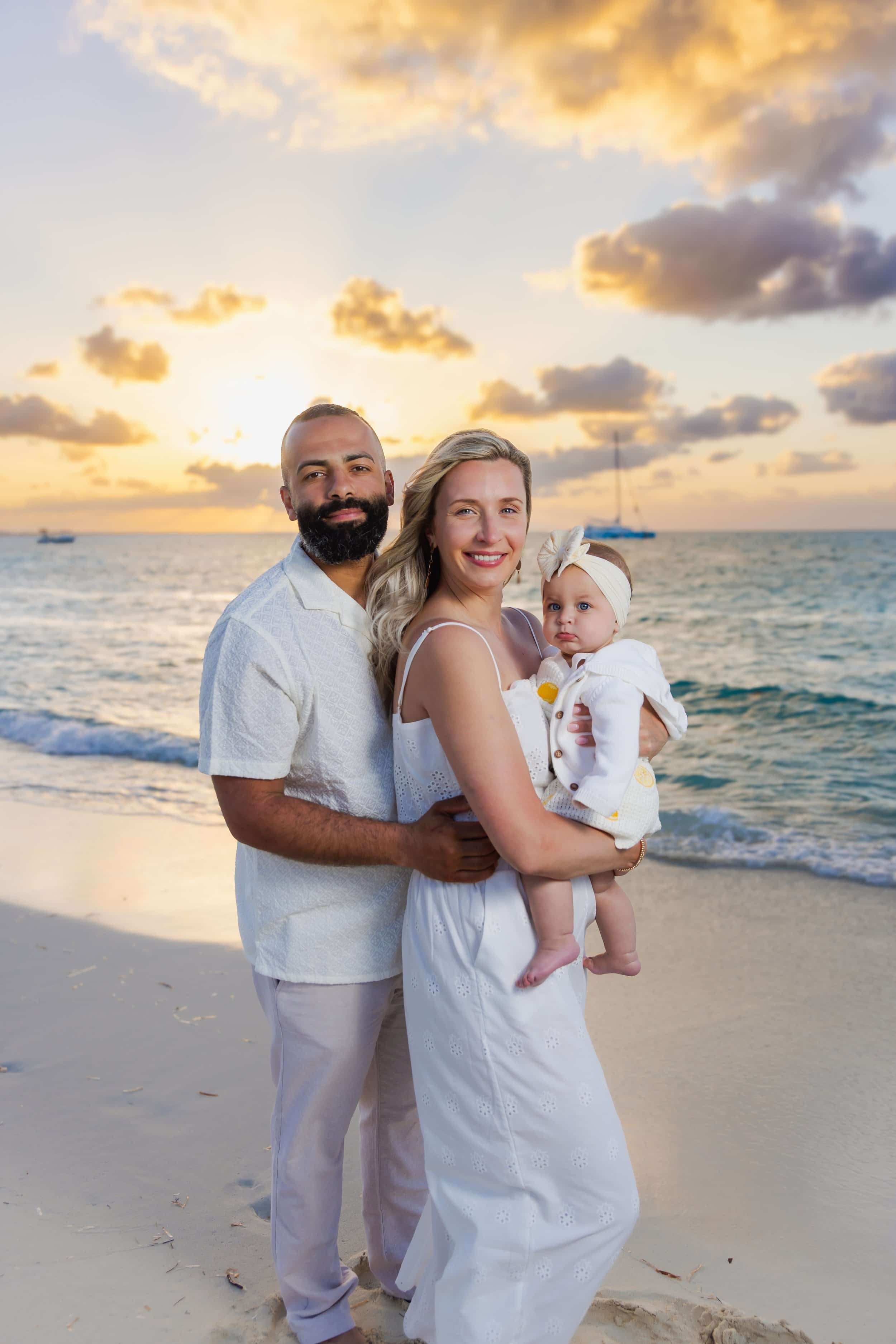 A family of three standing on the beach during sunset, with ocean waves and boats in the background. The man has a beard, the woman is holding a baby girl, and they are all dressed in white.