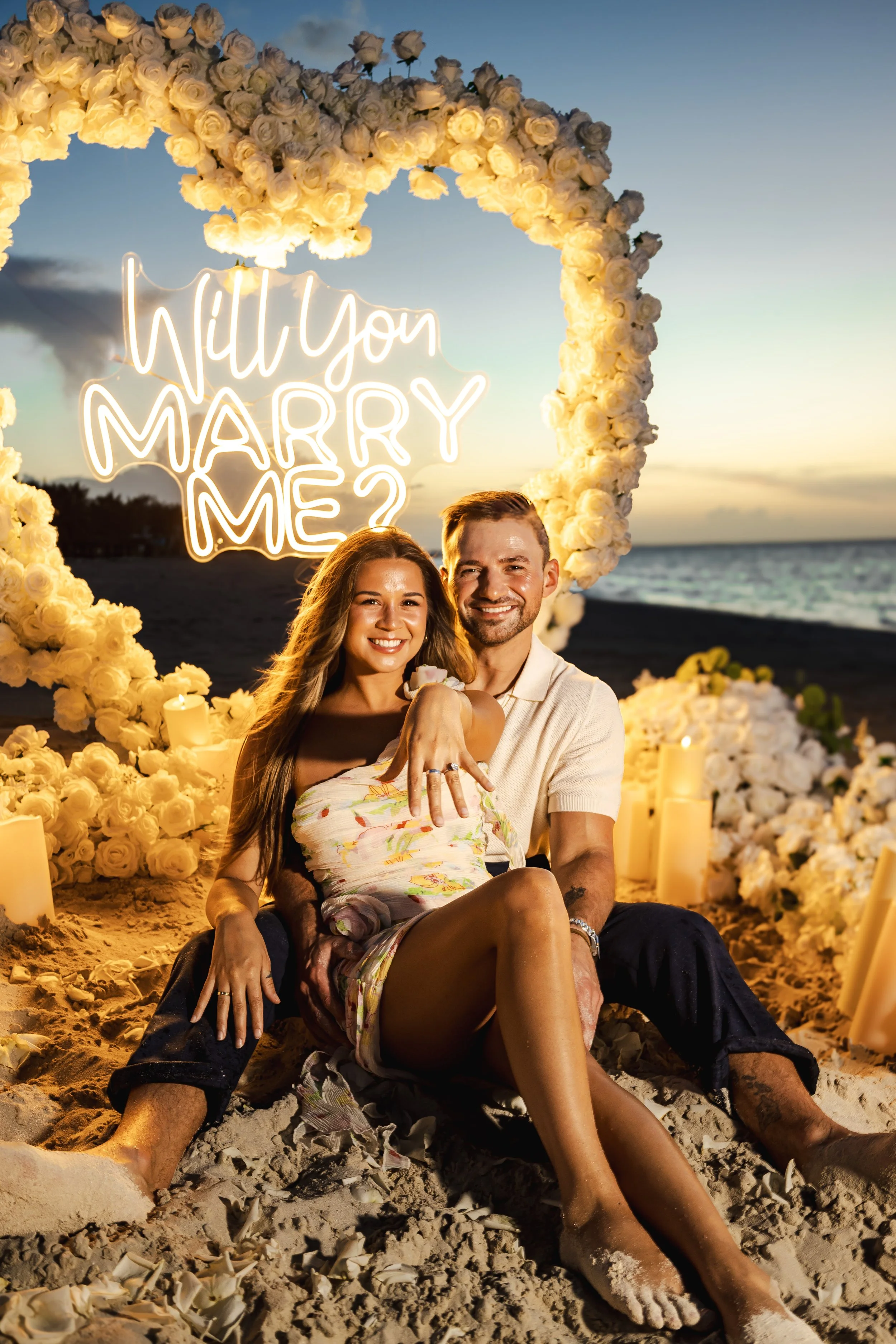 A couple sitting on the beach at sunset, surrounded by a floral heart-shaped arch with the neon sign 'Will You Marry Me?'. The woman is showing her engagement ring.