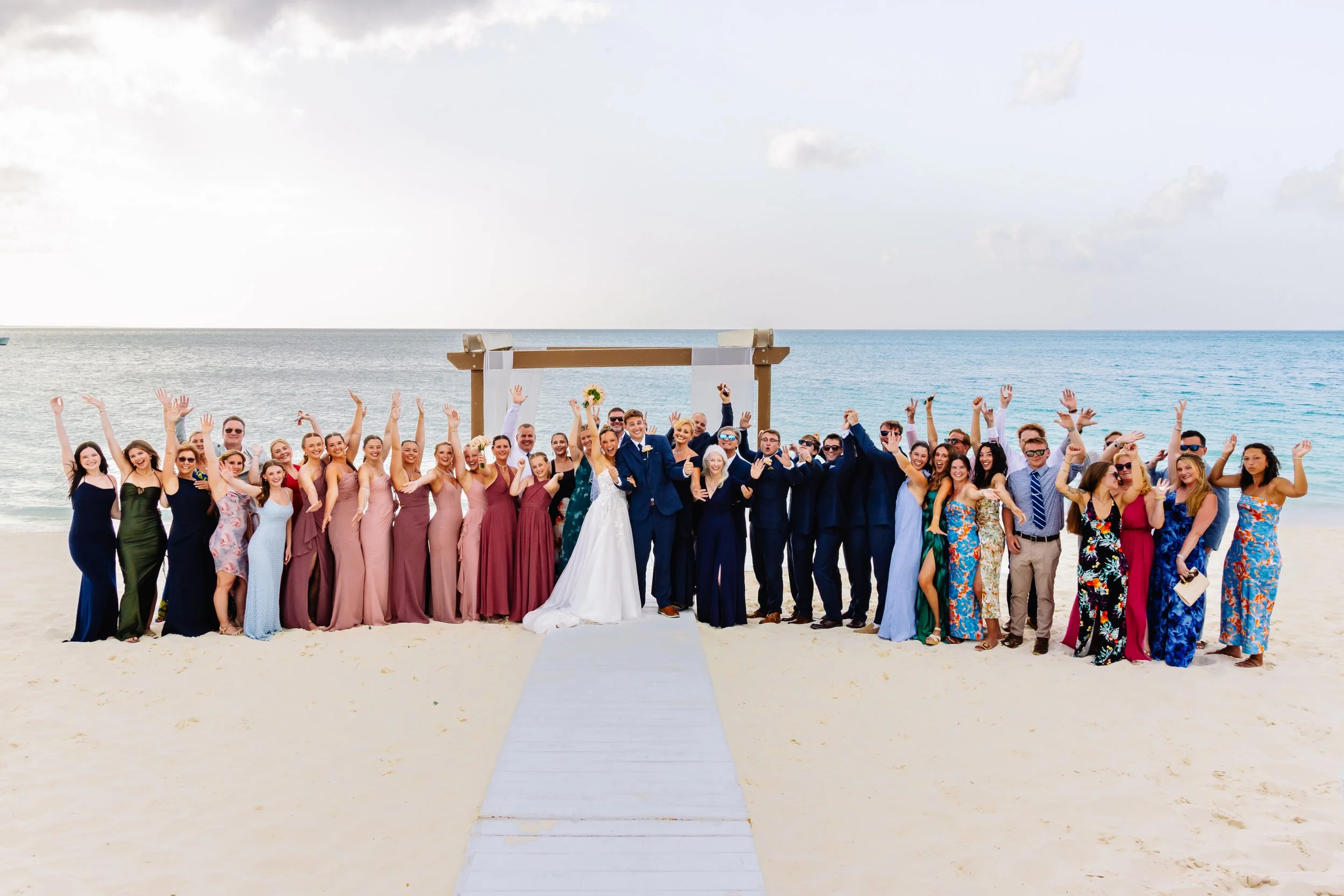 Group of wedding guests on the beach celebrating, with the bride and groom in the center, near an arch on the sandy shore with the ocean in the background.