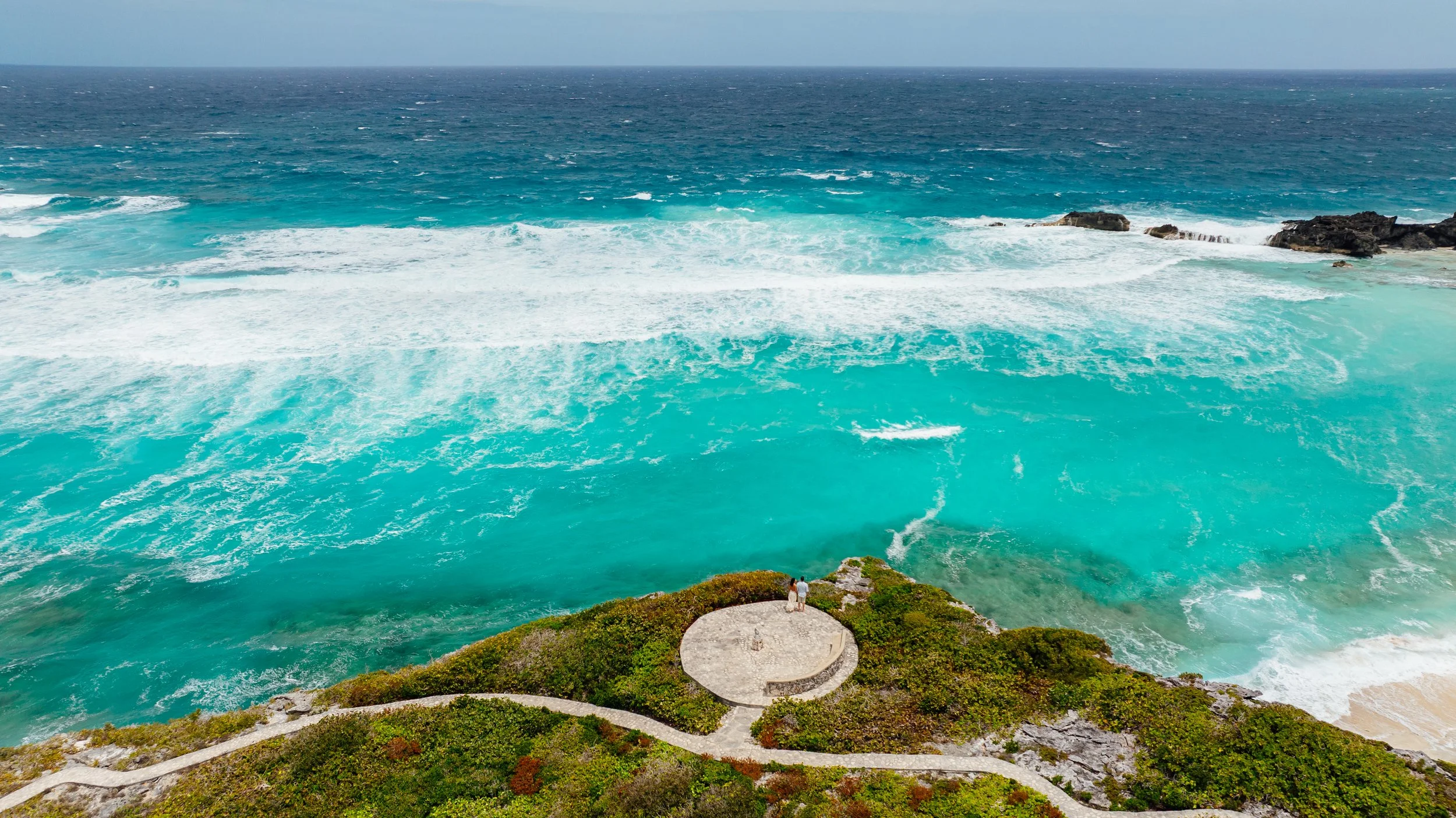 Two people standing on a circular platform overlooking the ocean with waves crashing against rocks and a rocky shoreline.