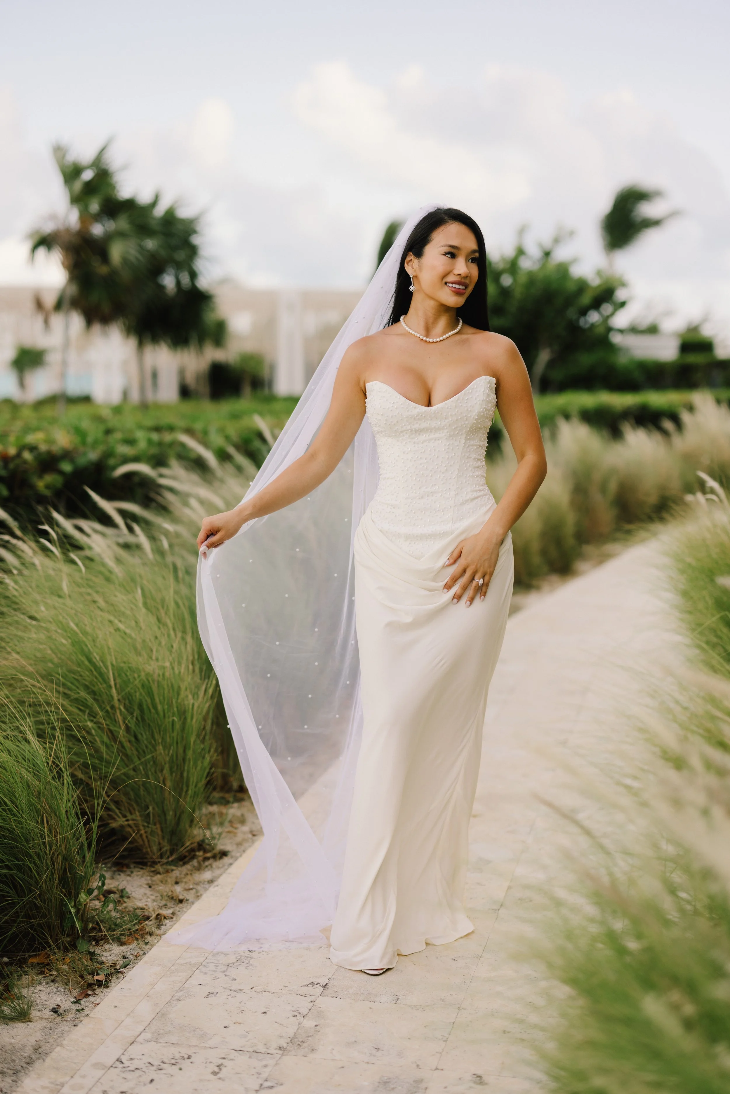 A woman in a white wedding dress with a veil and pearl necklace walking on a sandy path flanked by green plants and trees outside.
