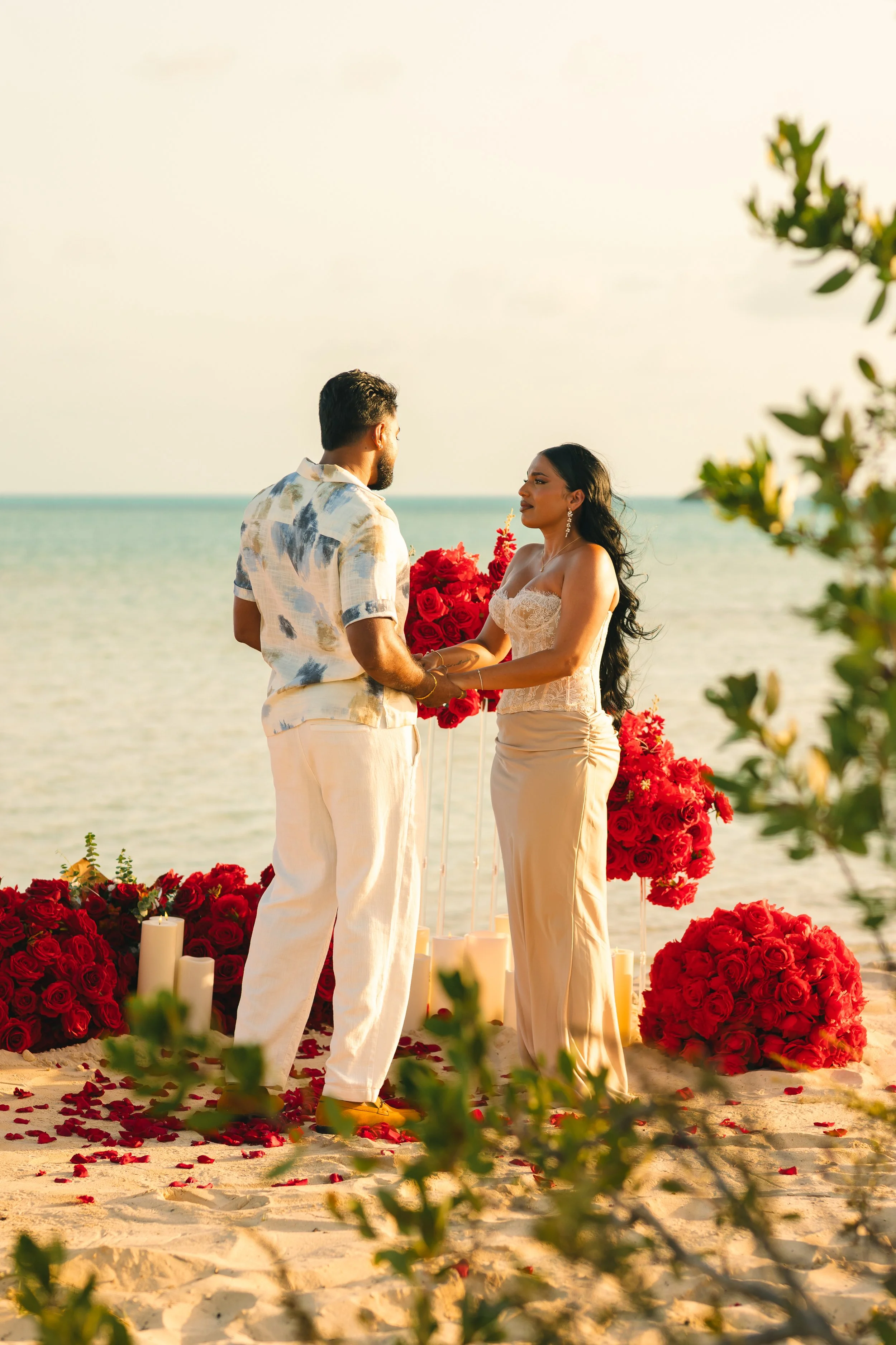A couple stands facing each other during a beach wedding ceremony, holding hands, with red roses and candles at their feet, the ocean and sky in the background.