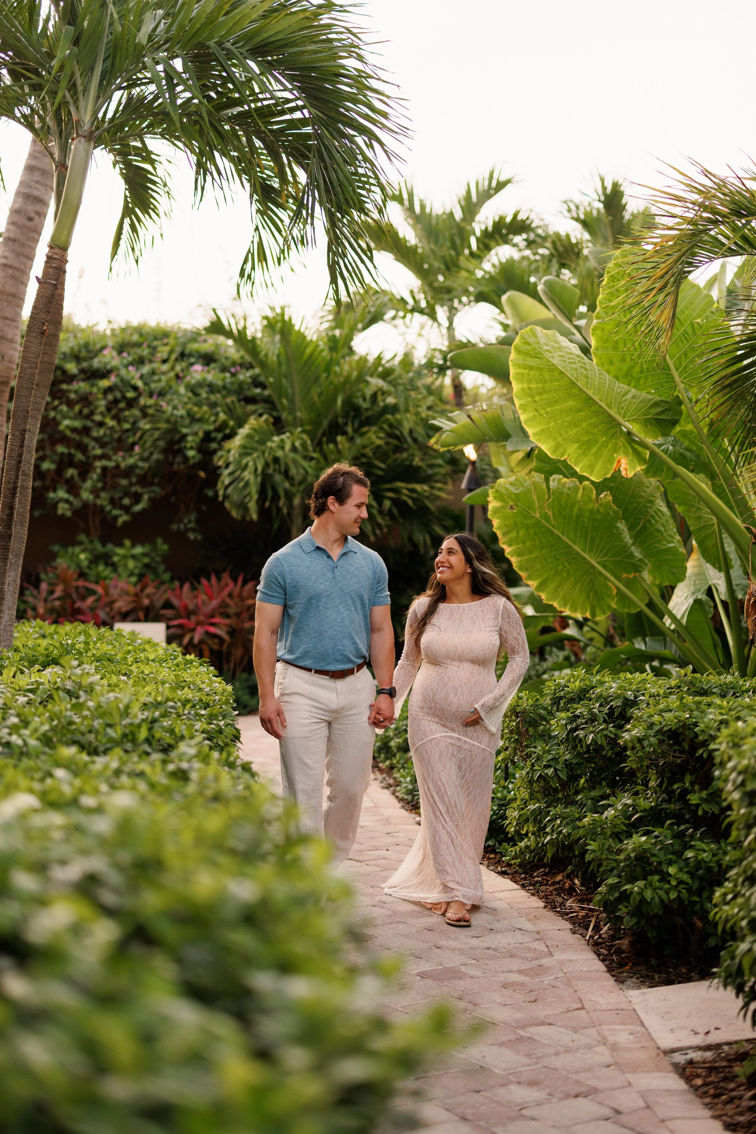 A couple walking hand in hand along a garden path surrounded by lush green tropical plants and trees, with the woman wearing a long lace dress and the man dressed in a blue shirt and khaki pants.