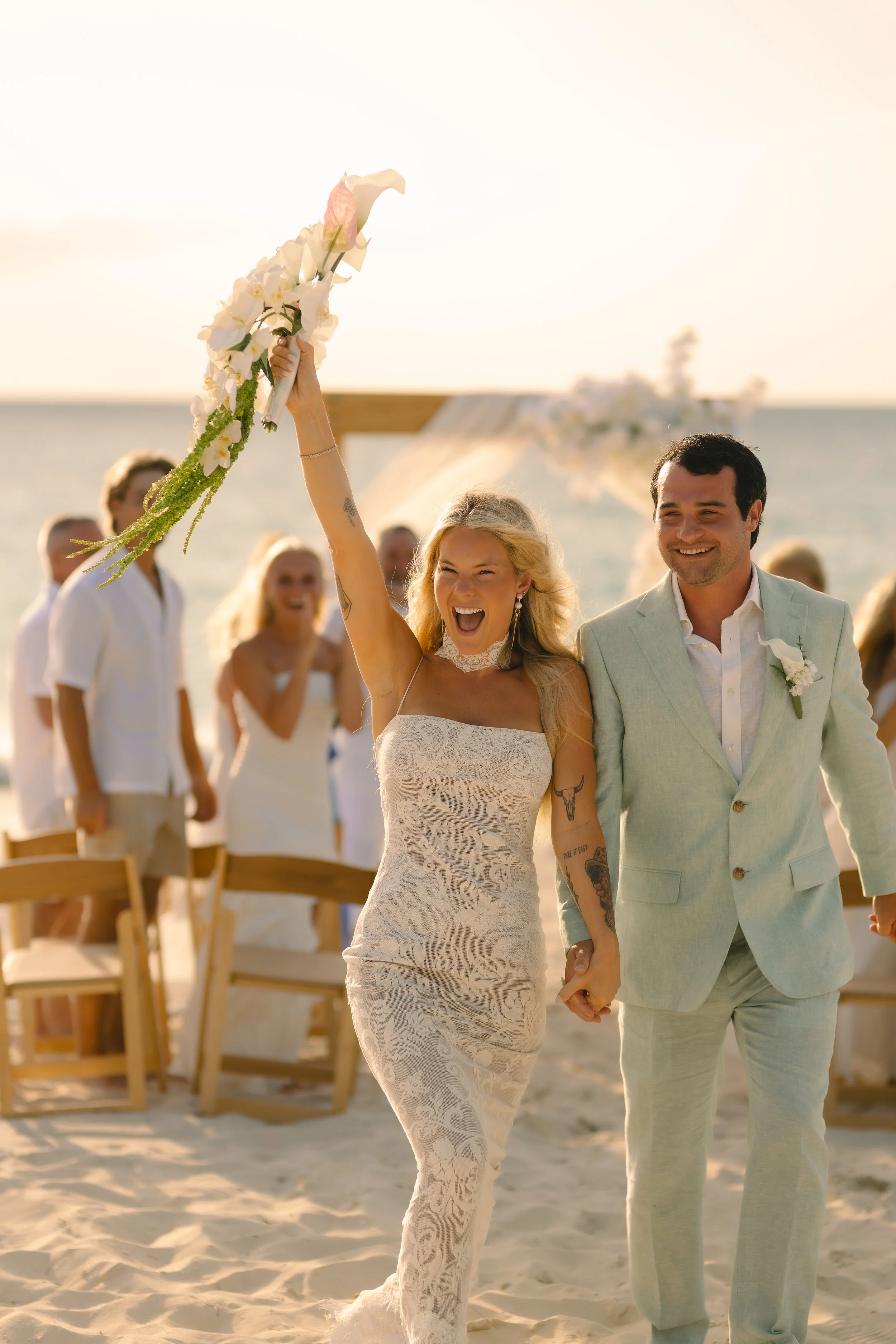 A bride and groom holding hands during their beach wedding ceremony at sunset, with guests in the background.