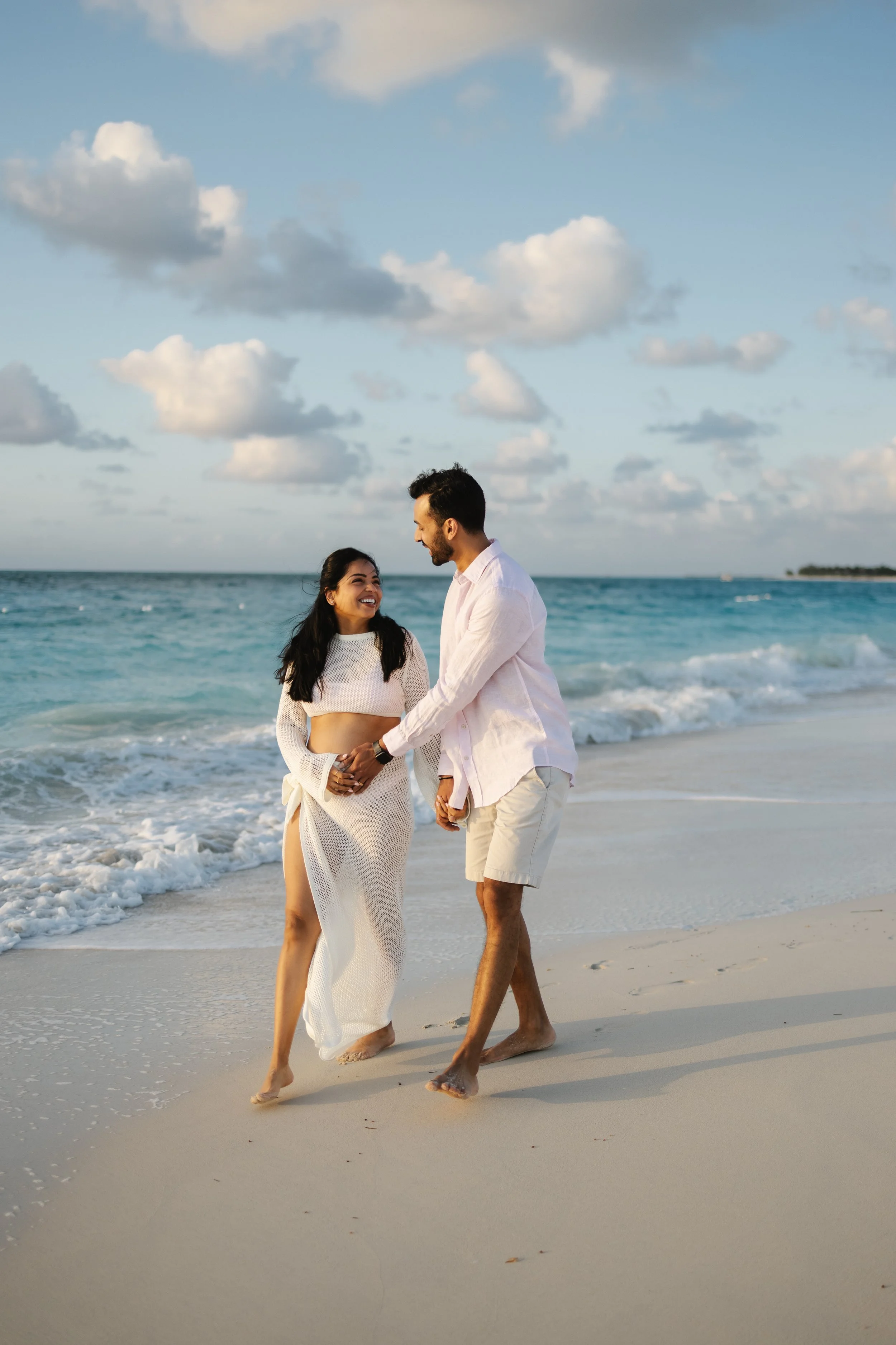 A couple holding hands and walking along the beach near the ocean, with clouds in the sky and a distant land on the horizon.