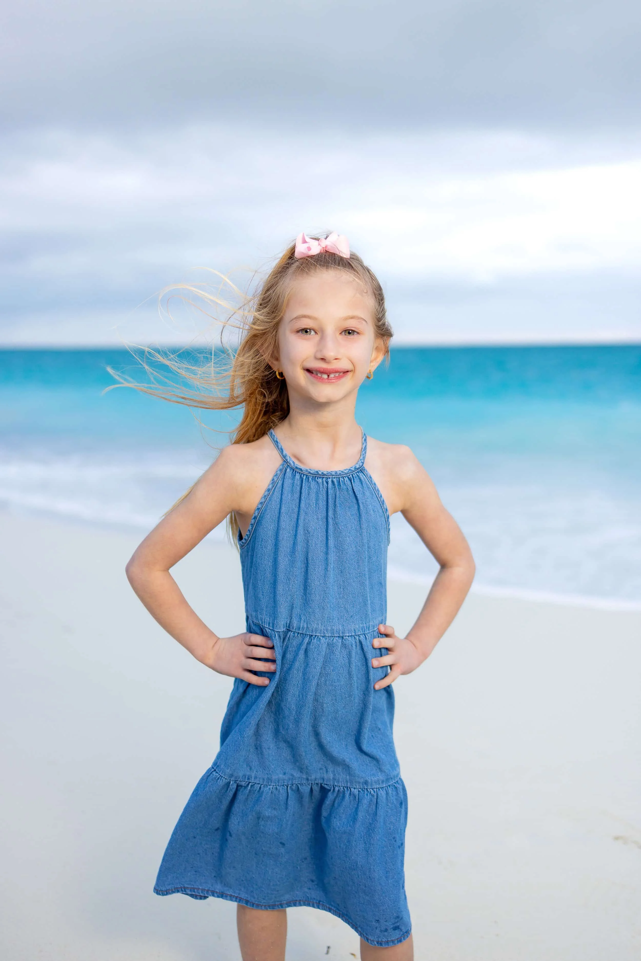 A young girl with blonde hair, wearing a blue denim dress and pink bow in her hair, standing on a beach with ocean waves and cloudy sky in the background.