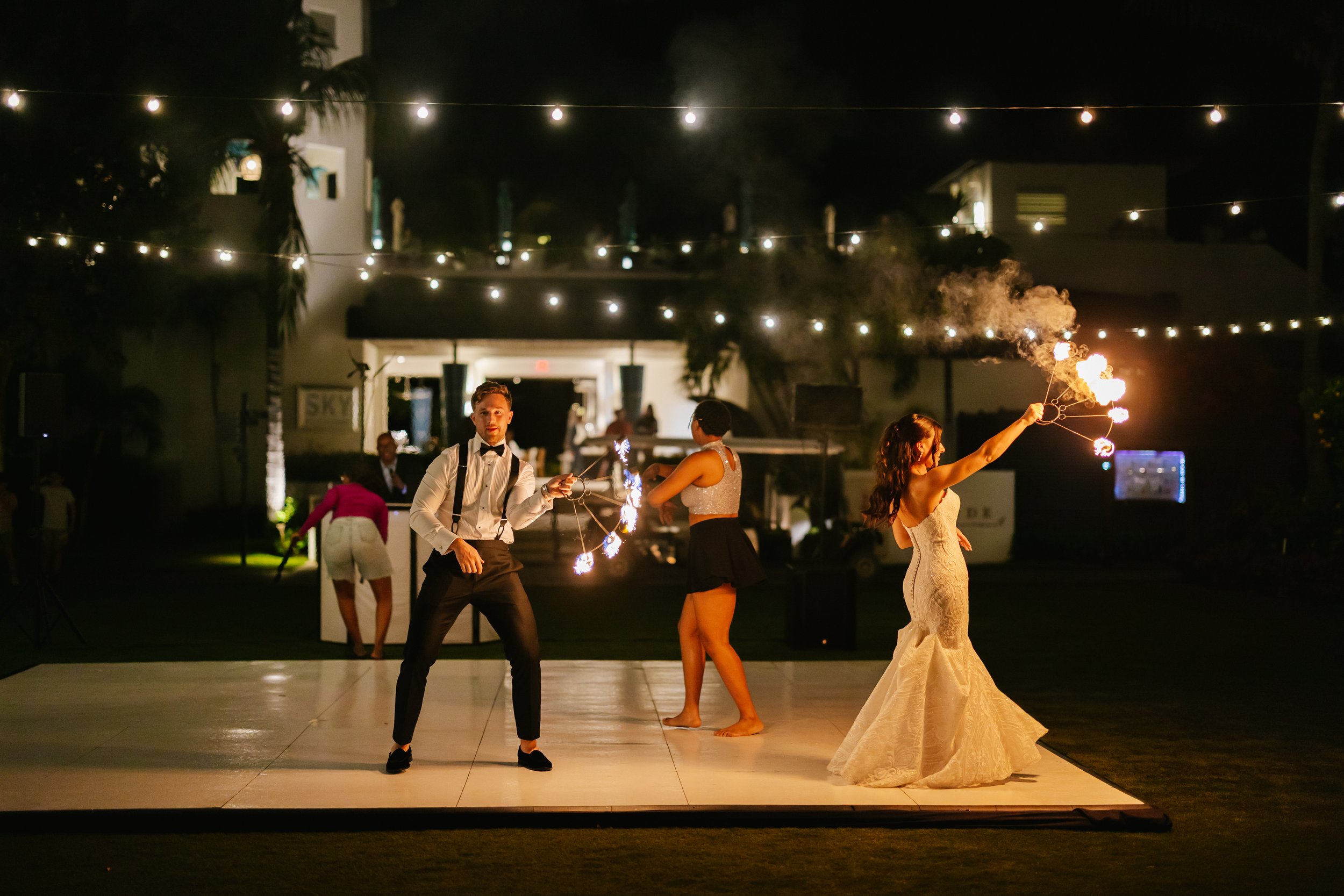 People performing a fire dance at night under string lights, with a woman in a wedding dress holding a fire poi.