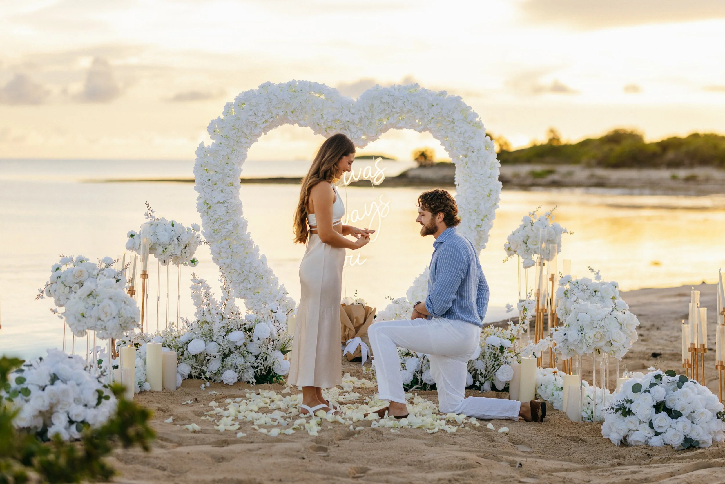 A couple is getting married on the beach during sunset, with a heart-shaped floral arch and arrangements of white flowers surrounding them.