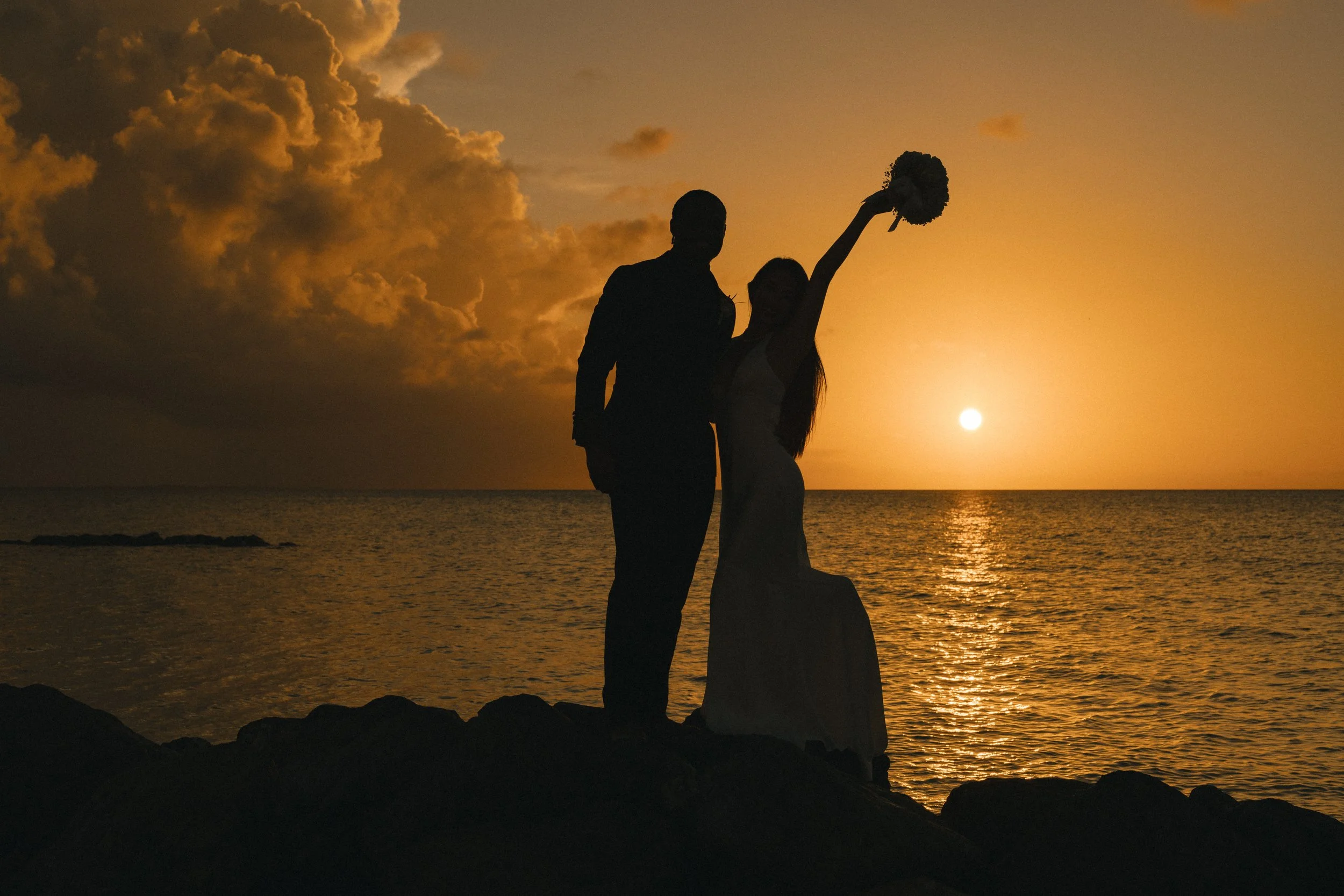 Silhouettes of a man and woman standing on rocks by the water during sunset, with the woman holding a bouquet of flowers in the air.