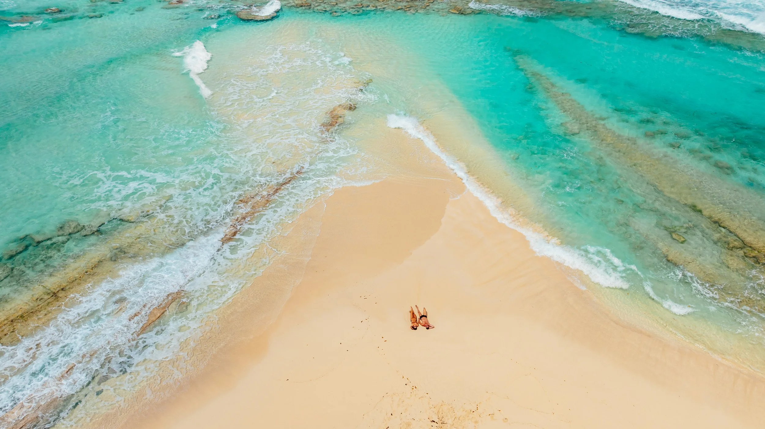 Two people lying on a sandy beach near clear turquoise water with gentle waves.