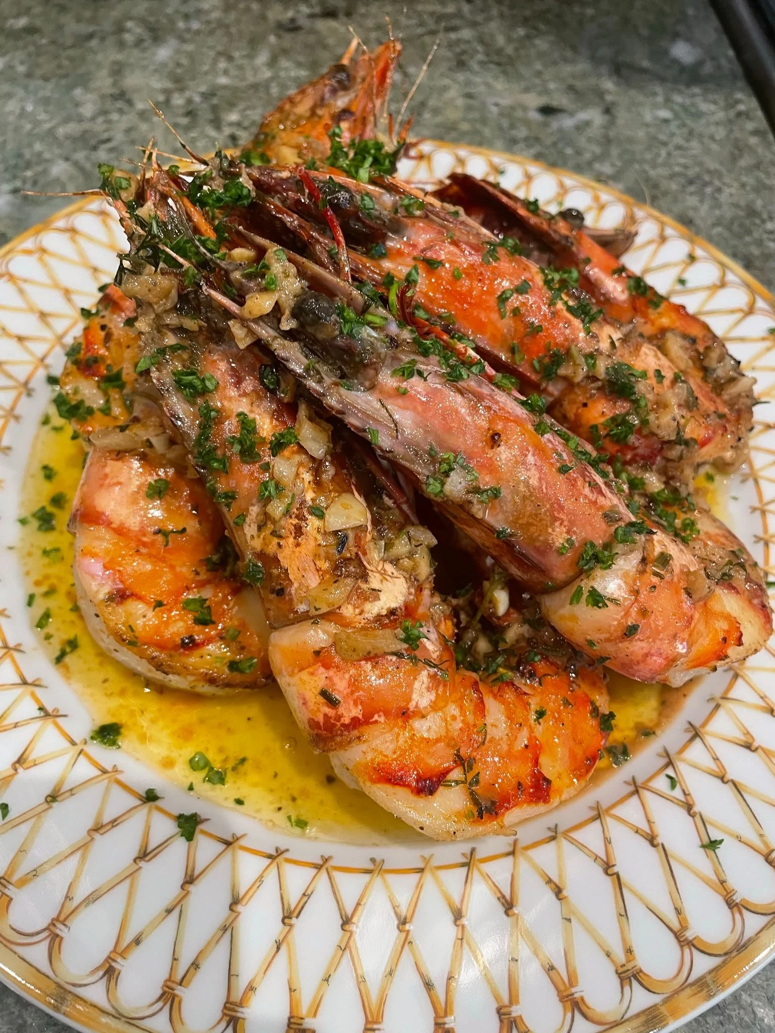 Close-up of cooked lobster tails garnished with chopped herbs on a decorative white plate with a gold geometric pattern, resting on a gray surface.