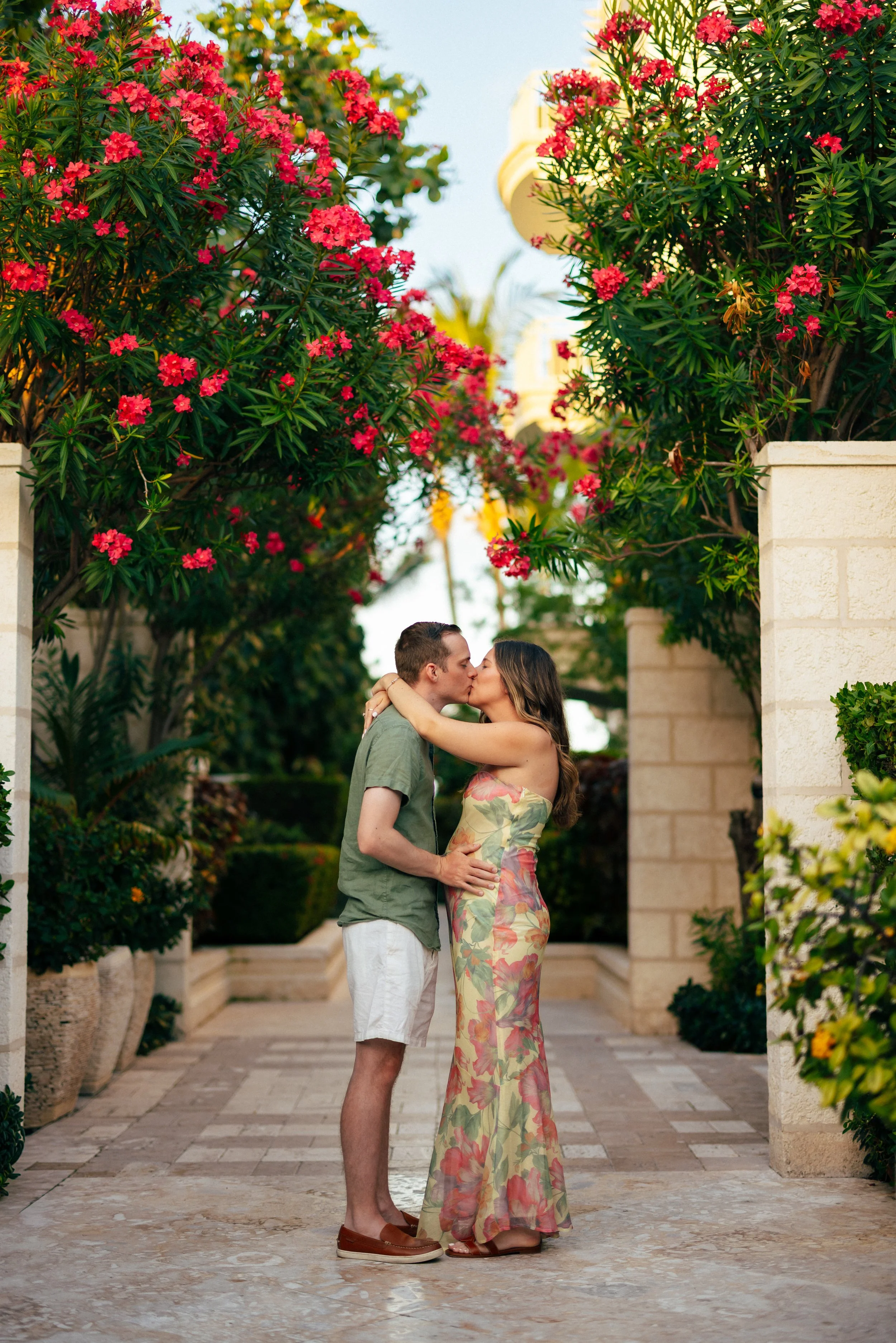 A couple kissing in a romantic embrace beneath pink flowering trees in an outdoor garden setting, with stone planters and a paved pathway.
