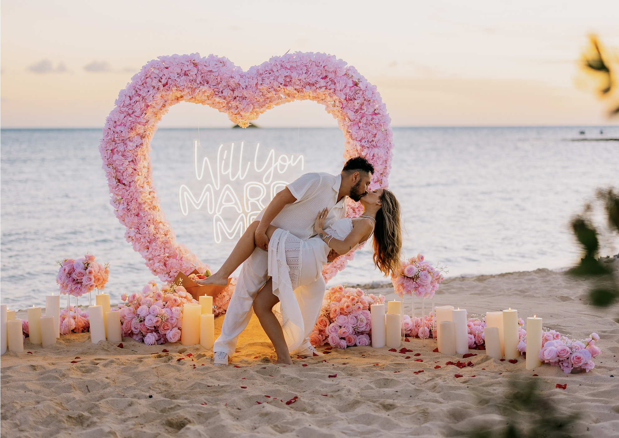 A couple sharing a kiss on a decorated beach setting with pink flowers, candles, and a heart-shaped floral frame with a sign that reads 'Will You Marry Me' in the background.