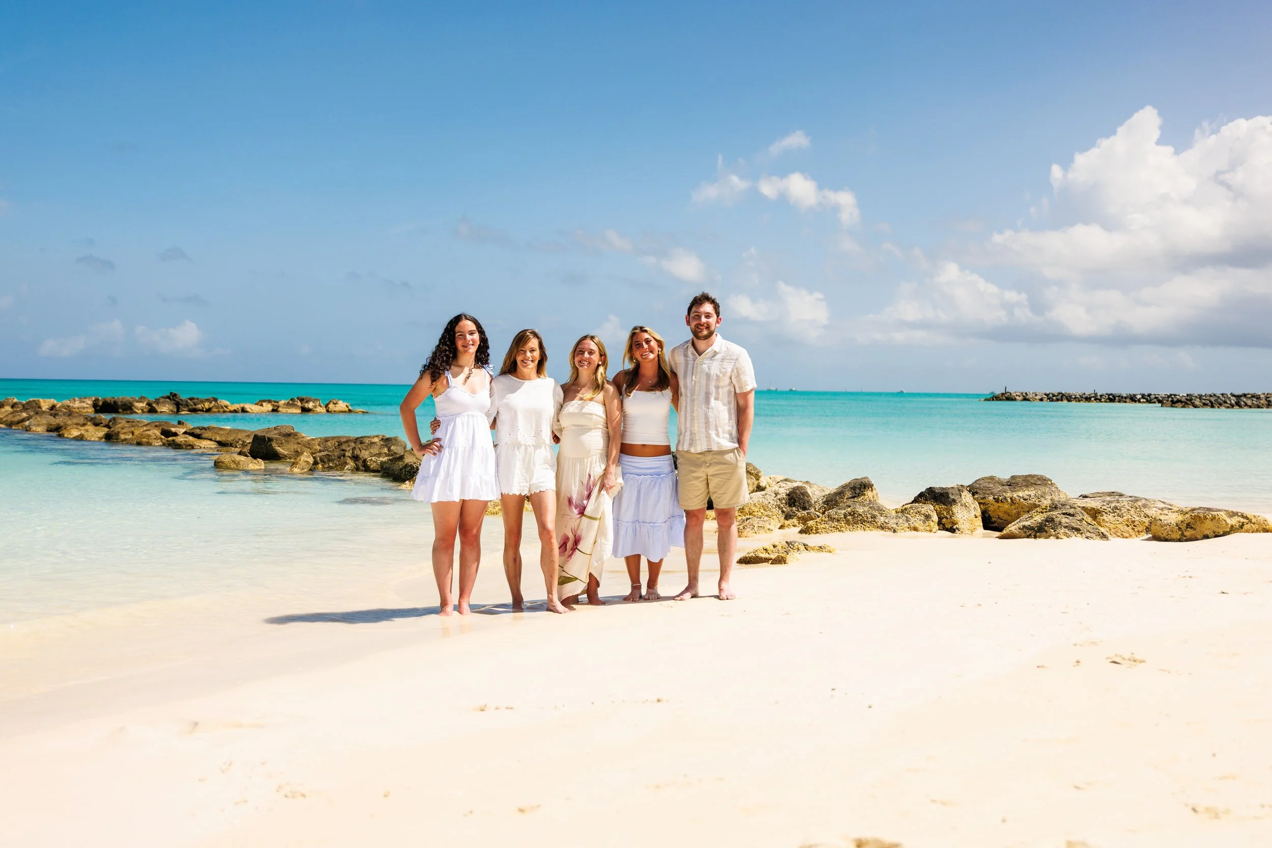 Group of five friends standing on a sandy beach near the ocean, smiling and posing for a photo, with rocks and the sea in the background under a partly cloudy sky.