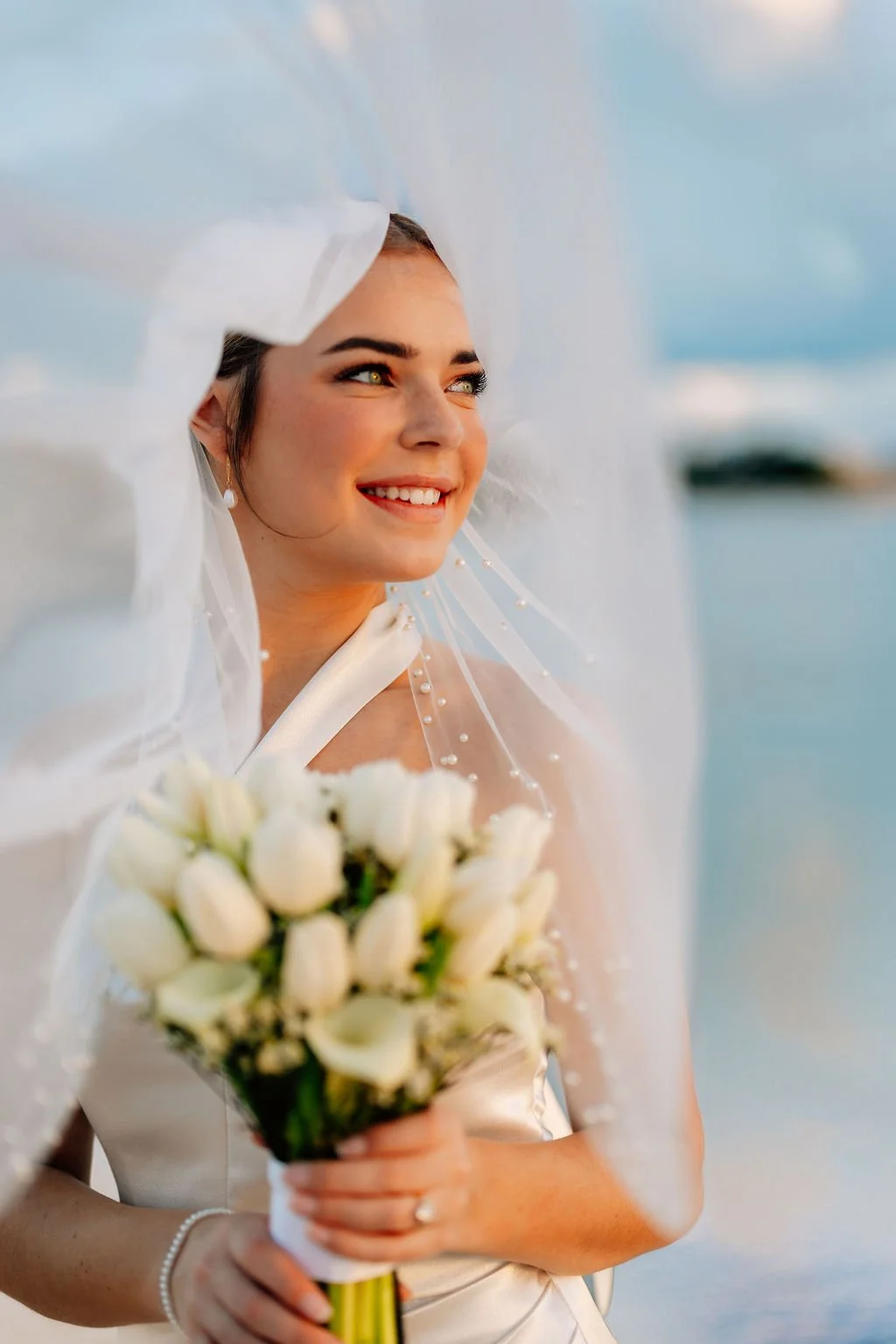 A bride on her wedding day, smiling and holding a bouquet of white flowers, with a veil partially covering her face, outdoors near water.