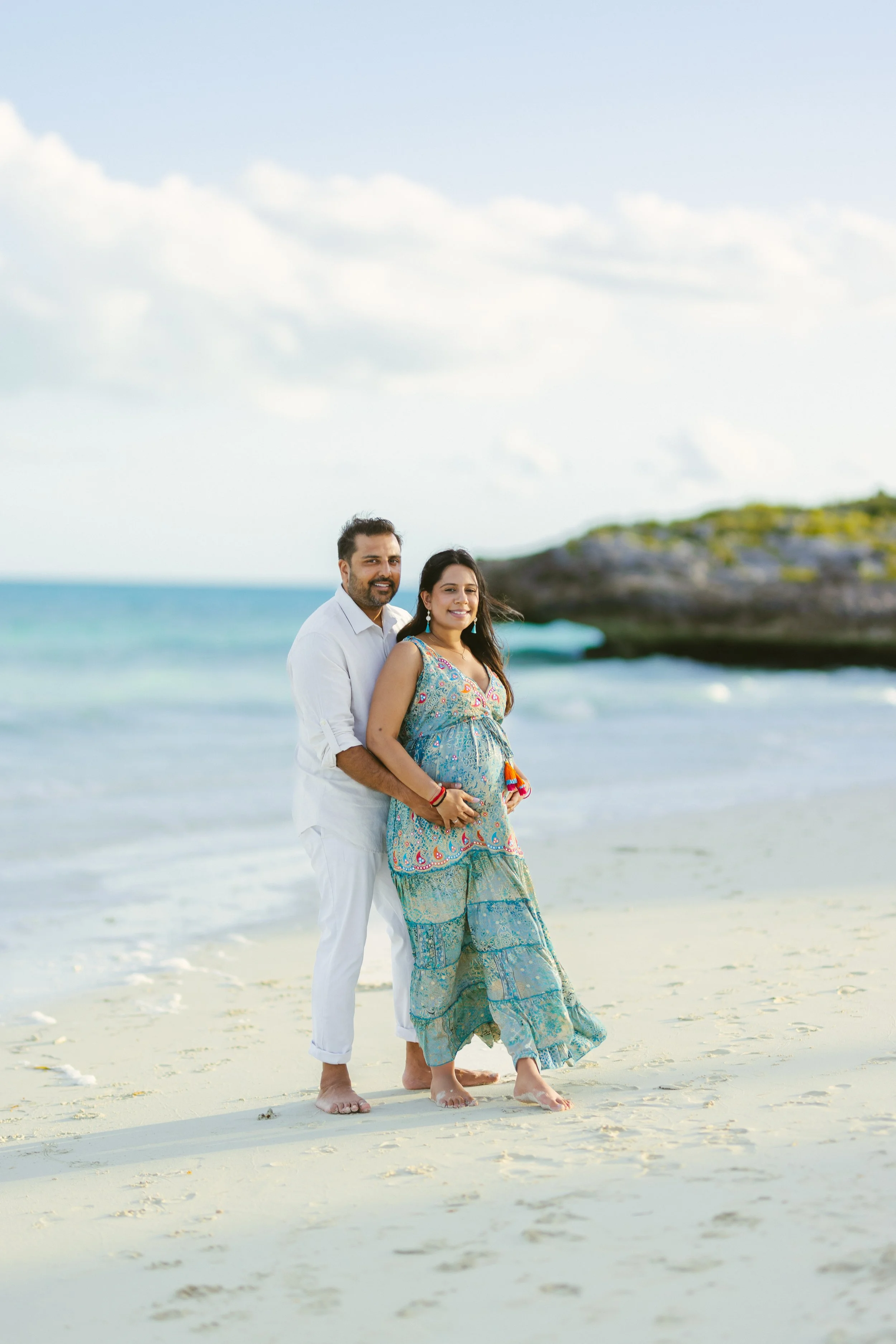 A couple standing on a sandy beach, with the ocean and a rocky headland in the background. The man is wearing white clothing, and the woman is wearing a colorful, patterned dress. They are both smiling and barefoot.