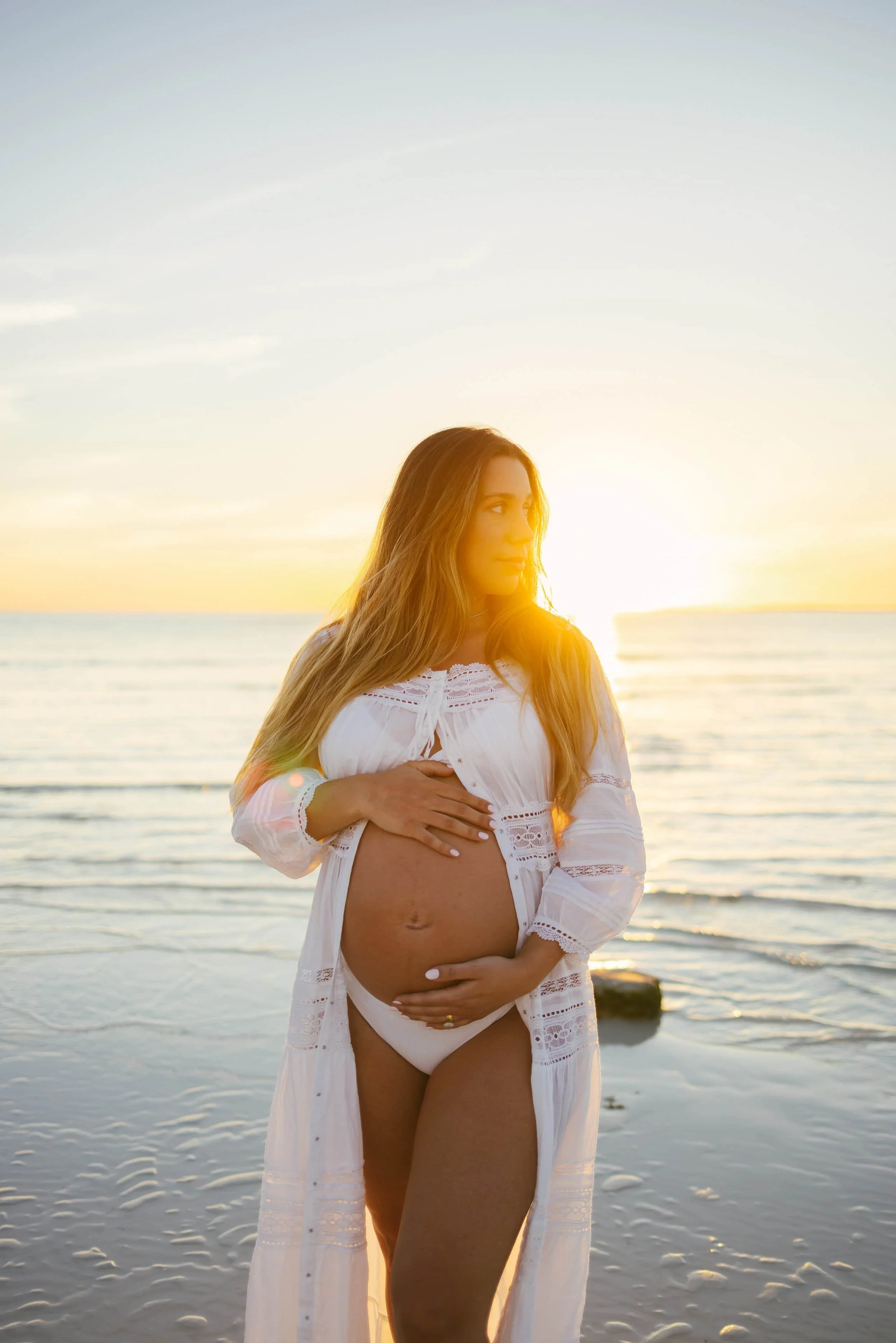 Pregnant woman in a white dress standing on the beach during sunset with her hand on her belly.