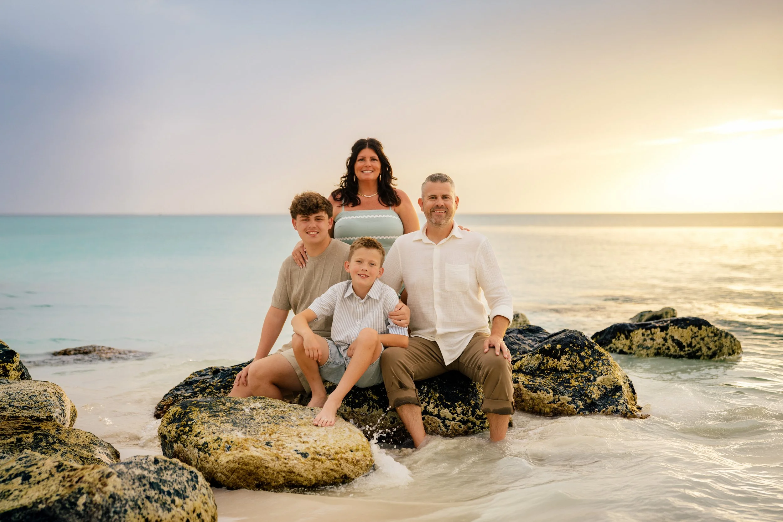A family of five sitting on rocks at the beach during sunset, smiling at the camera.