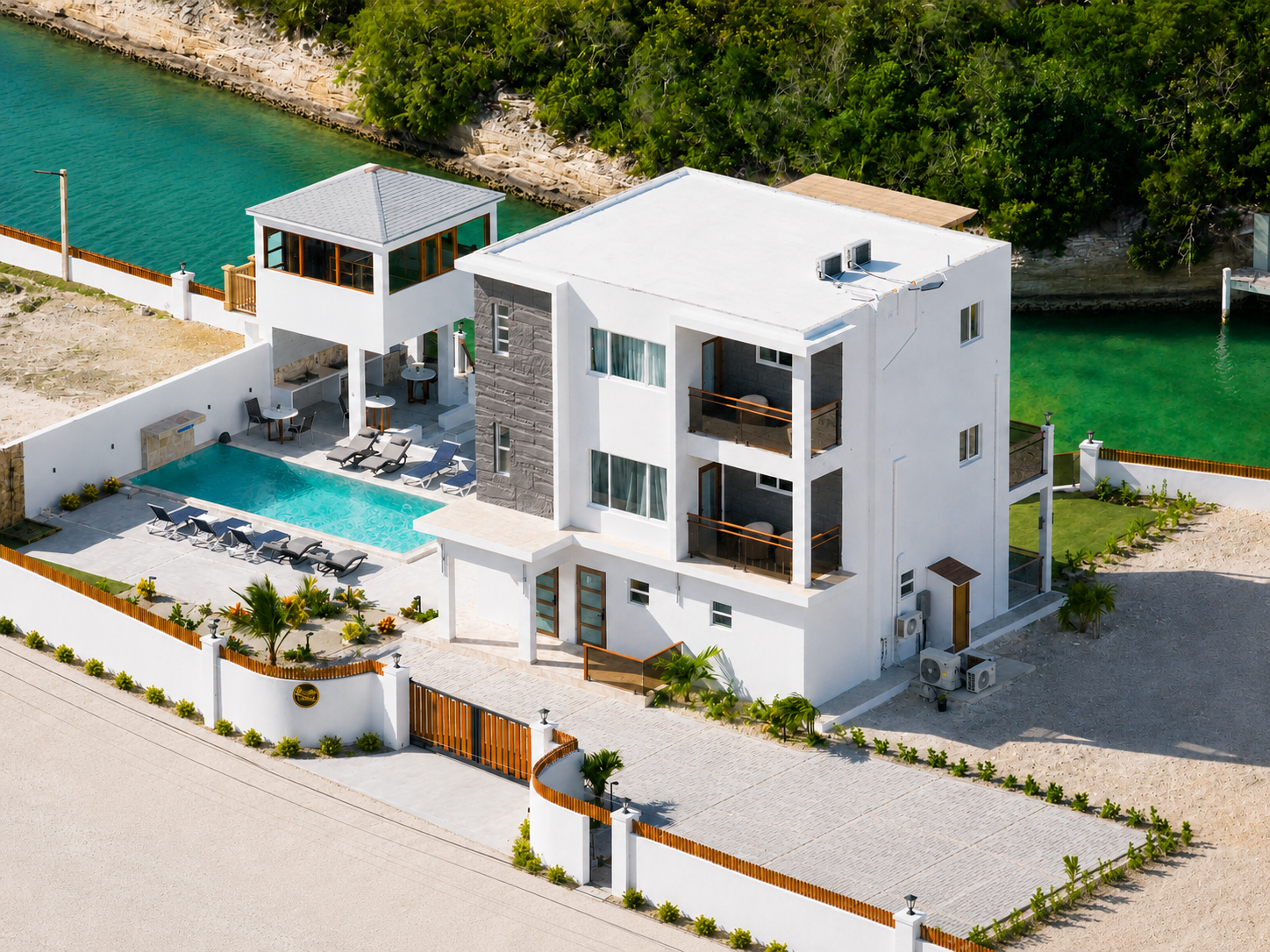 A modern white house with balconies, a swimming pool, outdoor lounge chairs, and a small house or gazebo nearby. The house is surrounded by a white fence and is situated by water, with trees in the background.