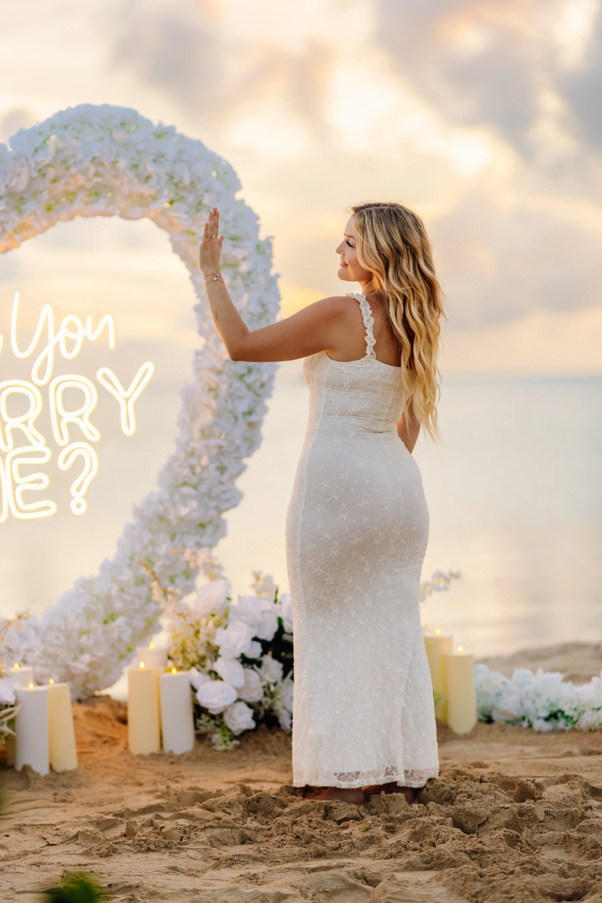 A woman in a white wedding dress standing on a sandy beach during sunset, with her hand raised in front of a floral arch and candles, against a cloudy sky backdrop.