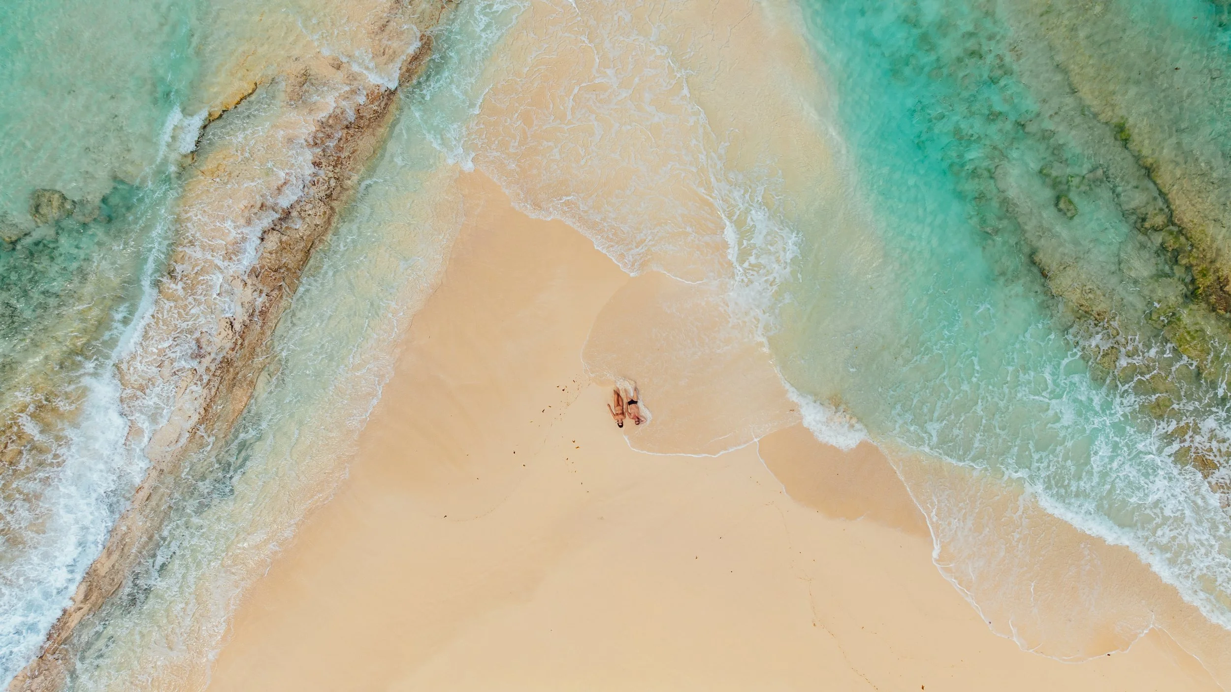 An aerial view of a sandy beach with turquoise waters and rocky formations, with two people lying on the sand near the water.