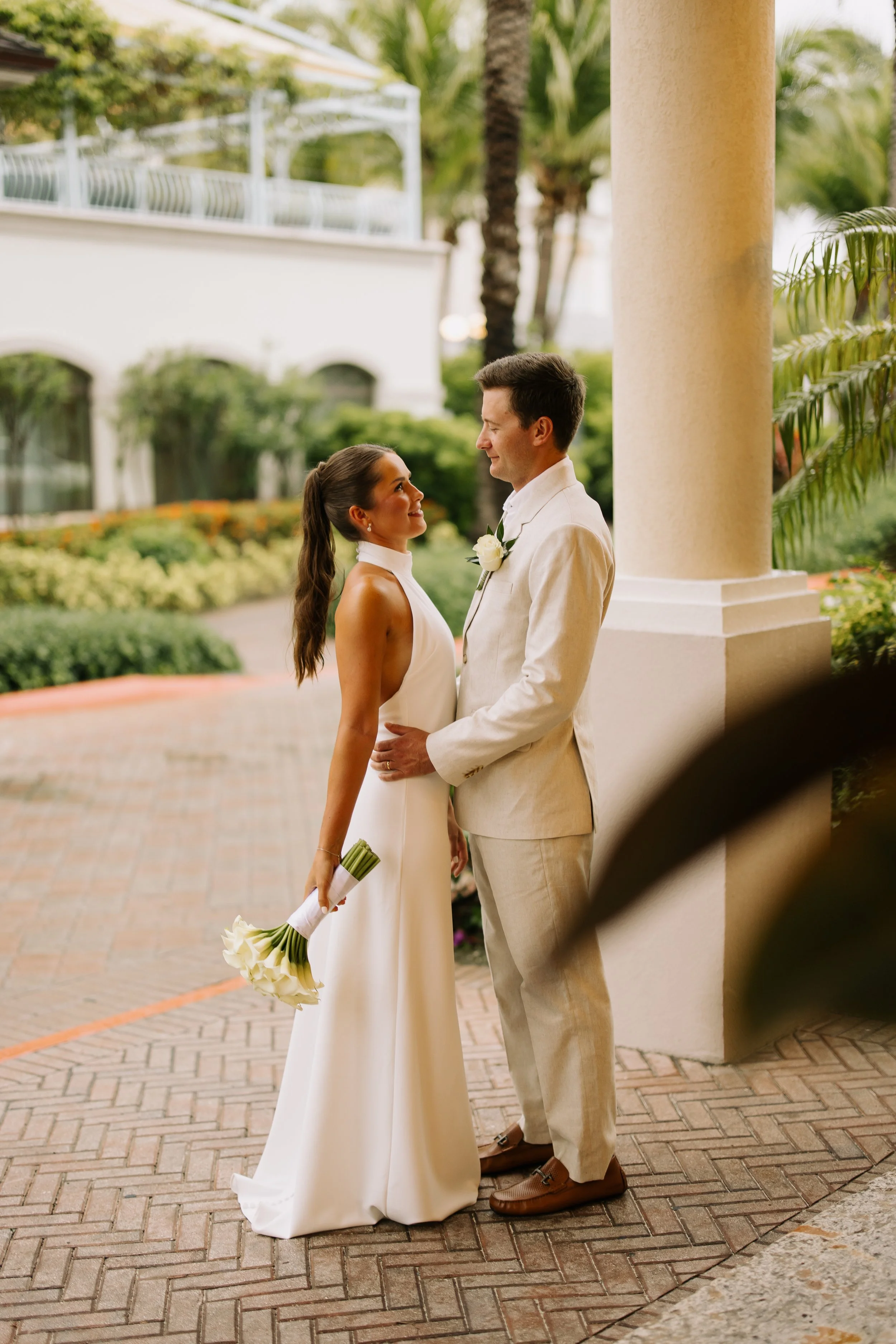 A bride and groom are looking at each other and smiling, standing outdoors under a column. The bride is holding a bouquet of white calla lilies, and the groom is wearing a light-colored suit with a white boutonniere. There is lush greenery and a whit