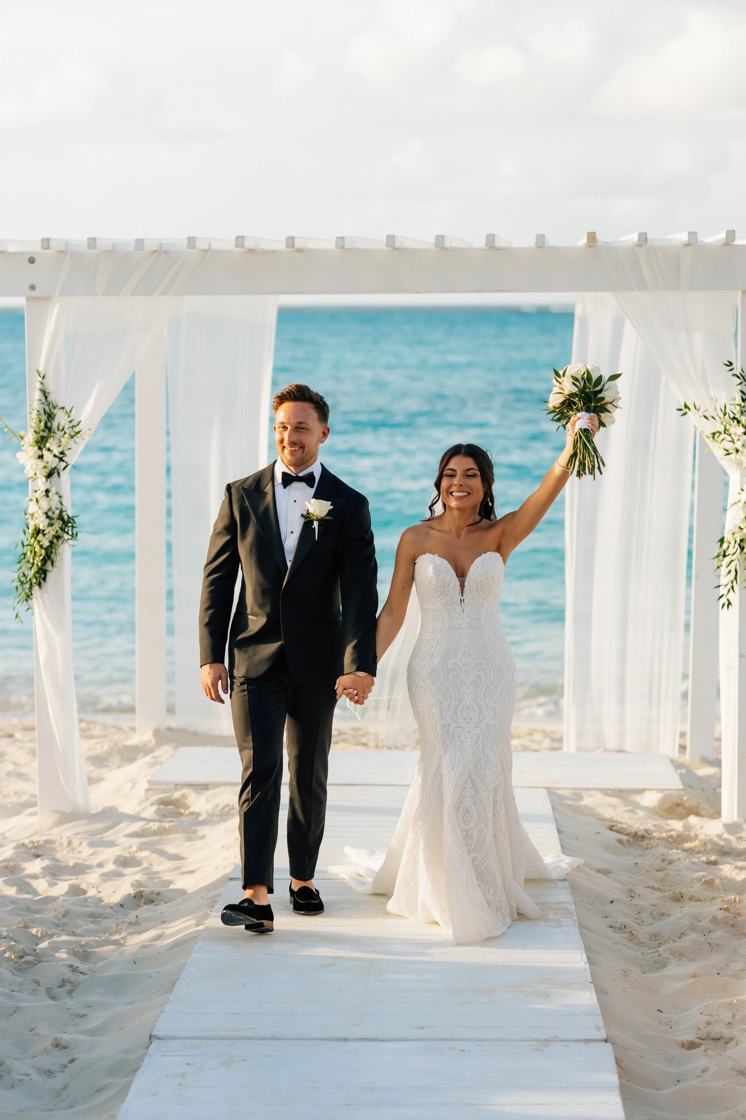 A bride and groom walking hand in hand on a beach under a wedding arch decorated with white fabric and flowers, with the ocean in the background.