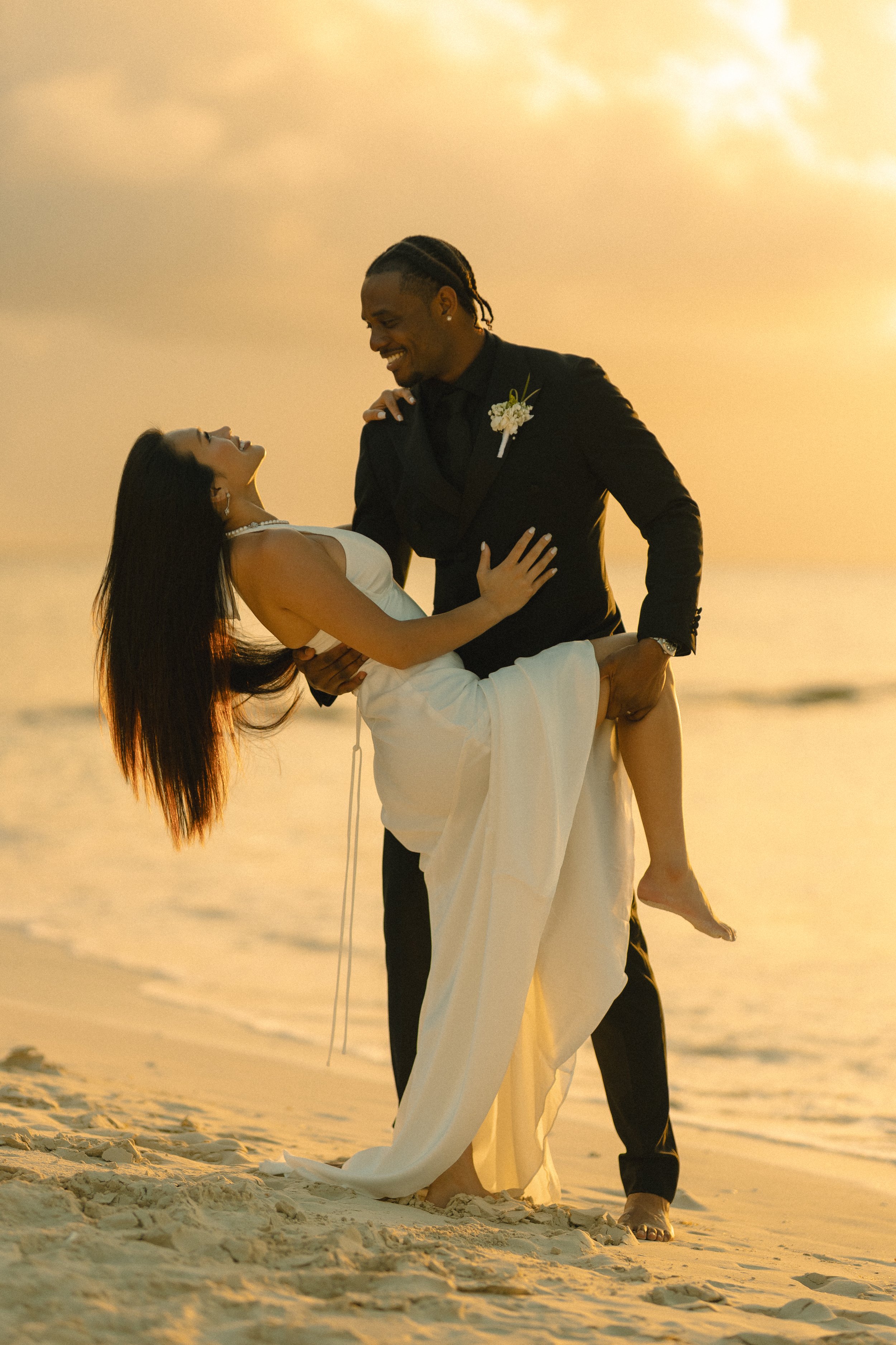 A wedding couple on the beach at sunset, with the groom holding the bride, who is smiling and looking up at him.