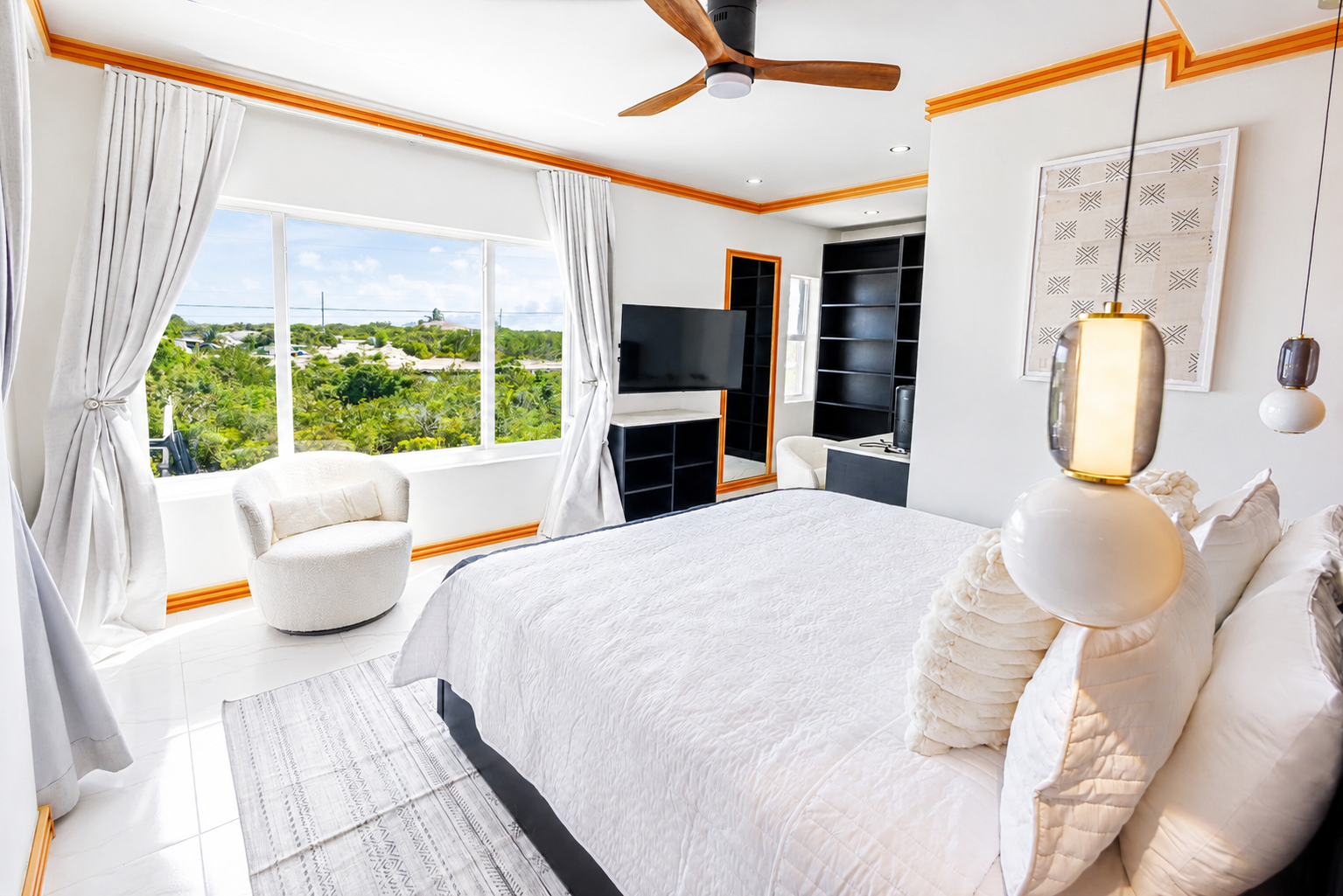 Bright bedroom with a large window providing a view of green trees outside, a white chair near the window, a television on a black stand, a black bookshelf, a bed with white bedding and pillows, and modern hanging lamps.