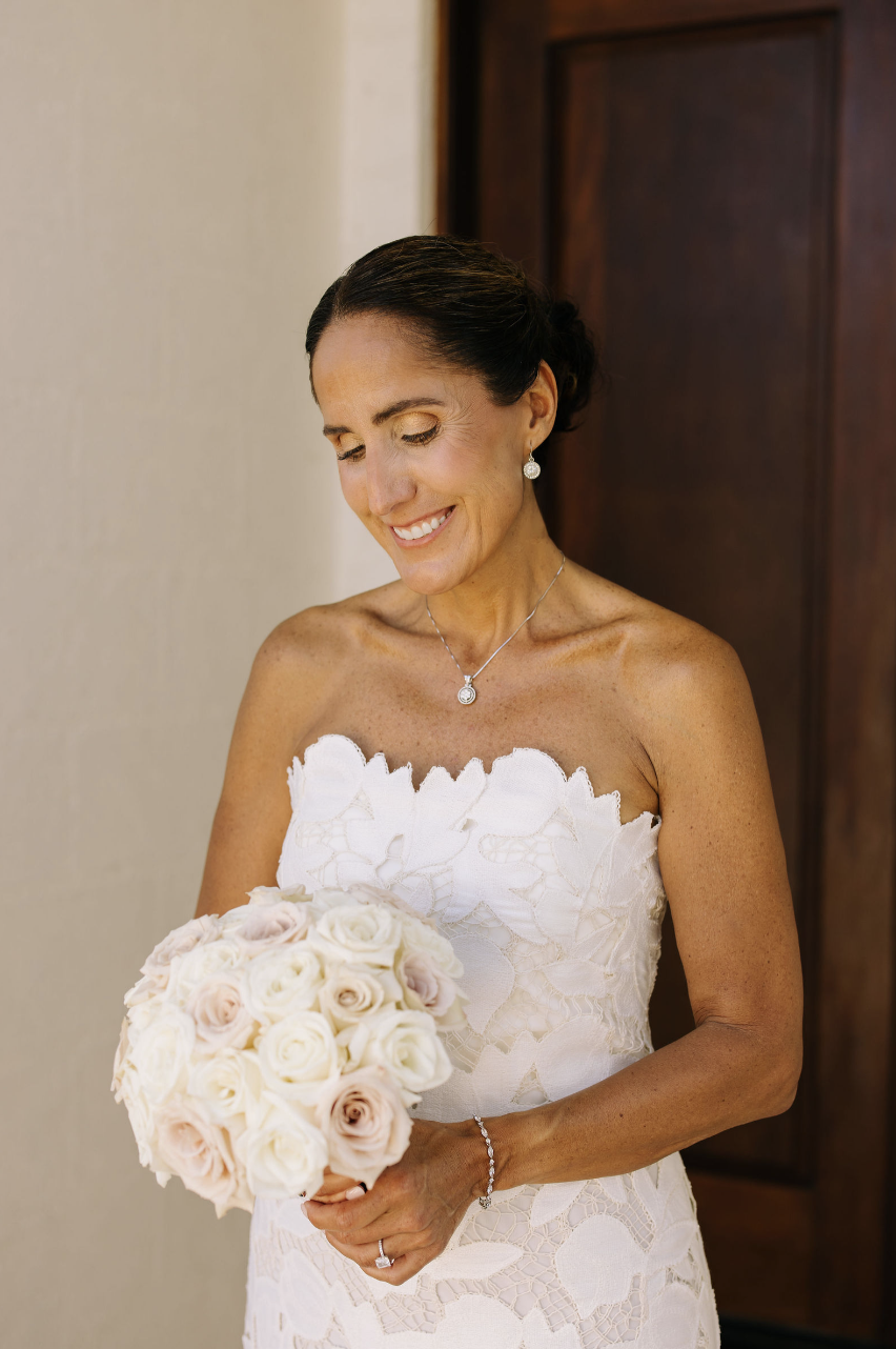 A smiling bride in a white lace strapless wedding dress holding a bouquet of white and blush roses.