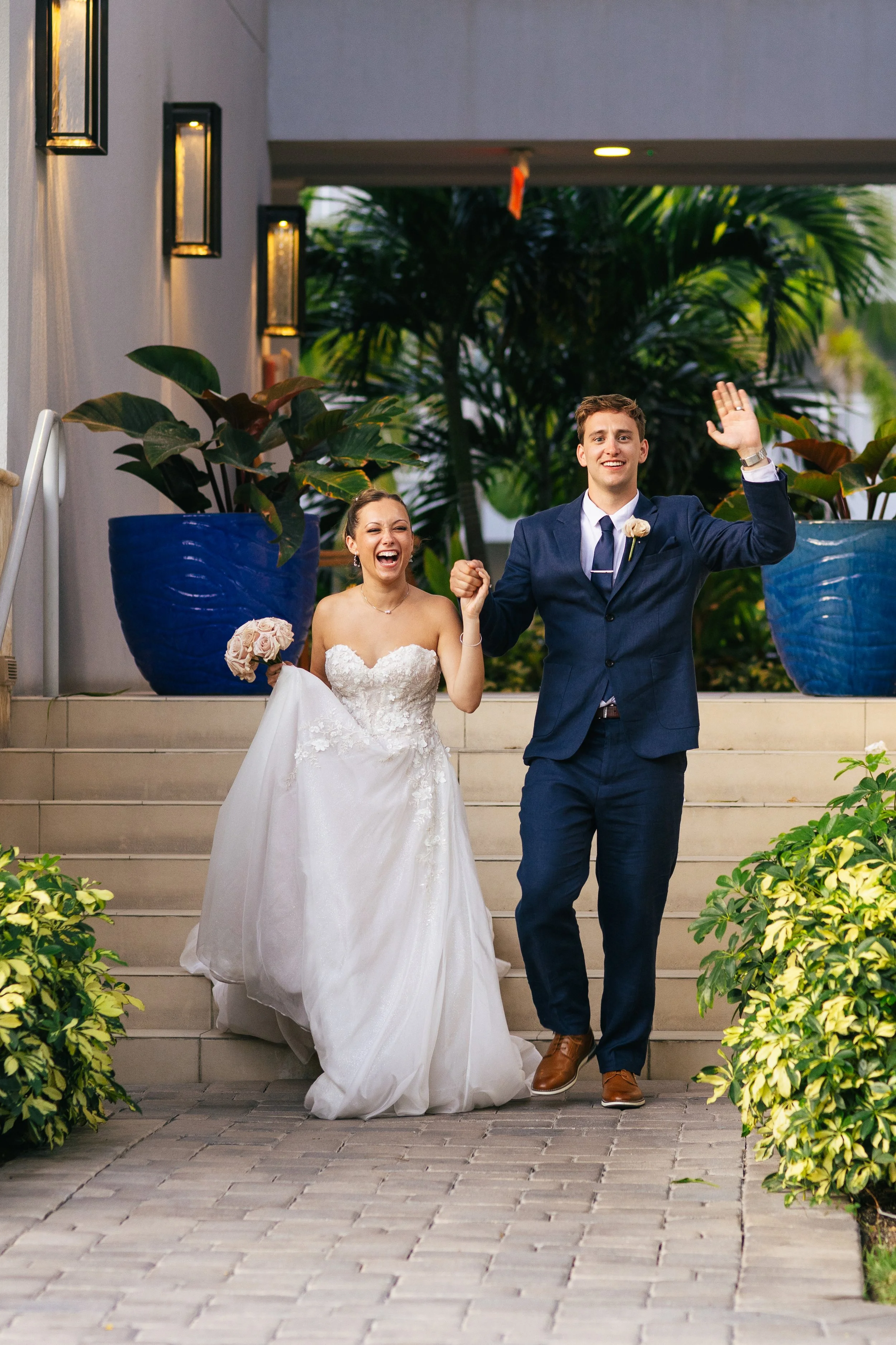 A newlywed couple walking down stairs, the bride wearing a white wedding gown and holding a bouquet, the groom in a dark blue suit raising his hand, both appearing joyful and happy to celebrate their wedding.
