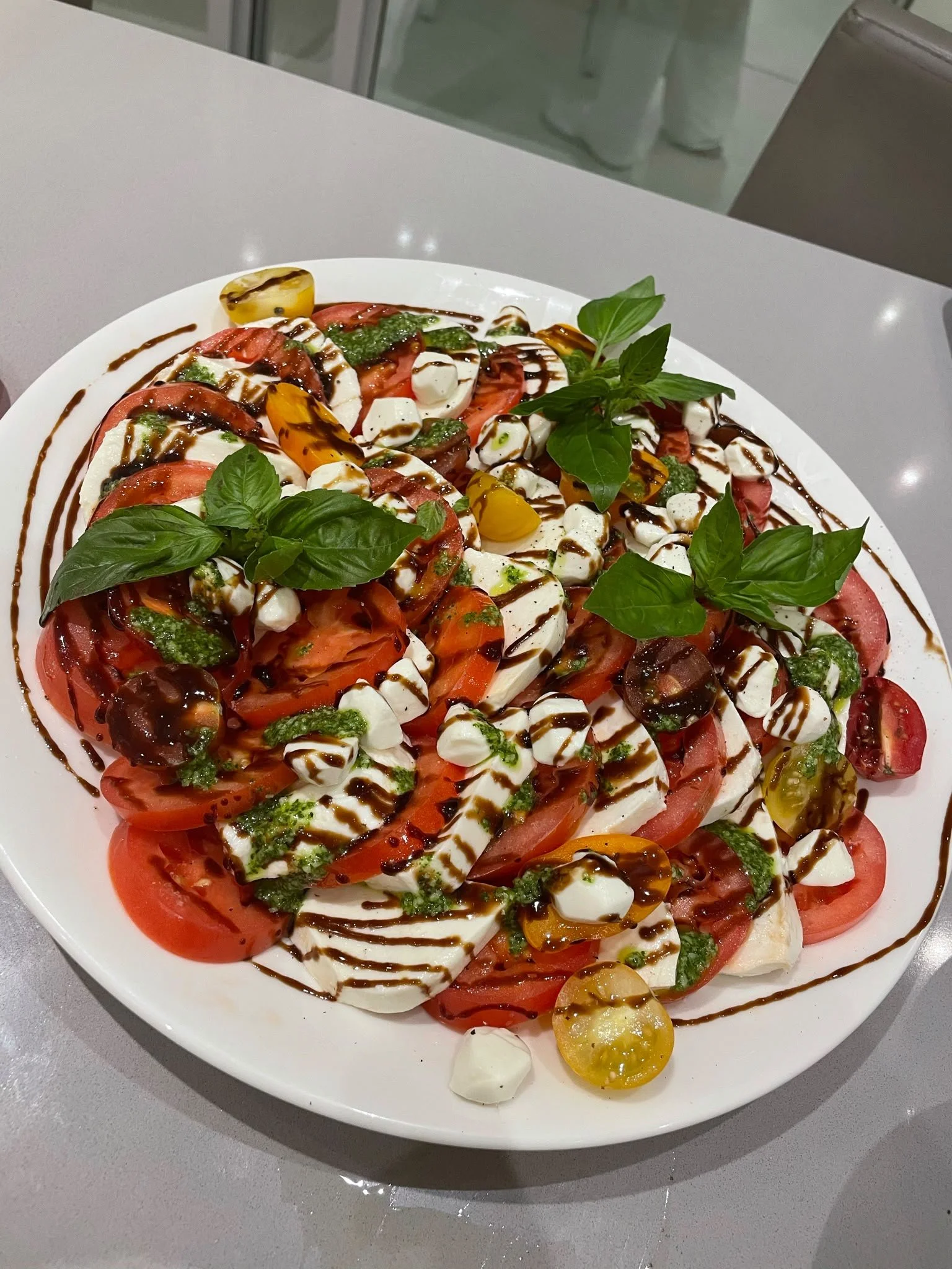 Caprese salad with sliced tomatoes, mozzarella cheese, basil leaves, cherry tomatoes, drizzled with balsamic glaze and green pesto, served on a white plate.