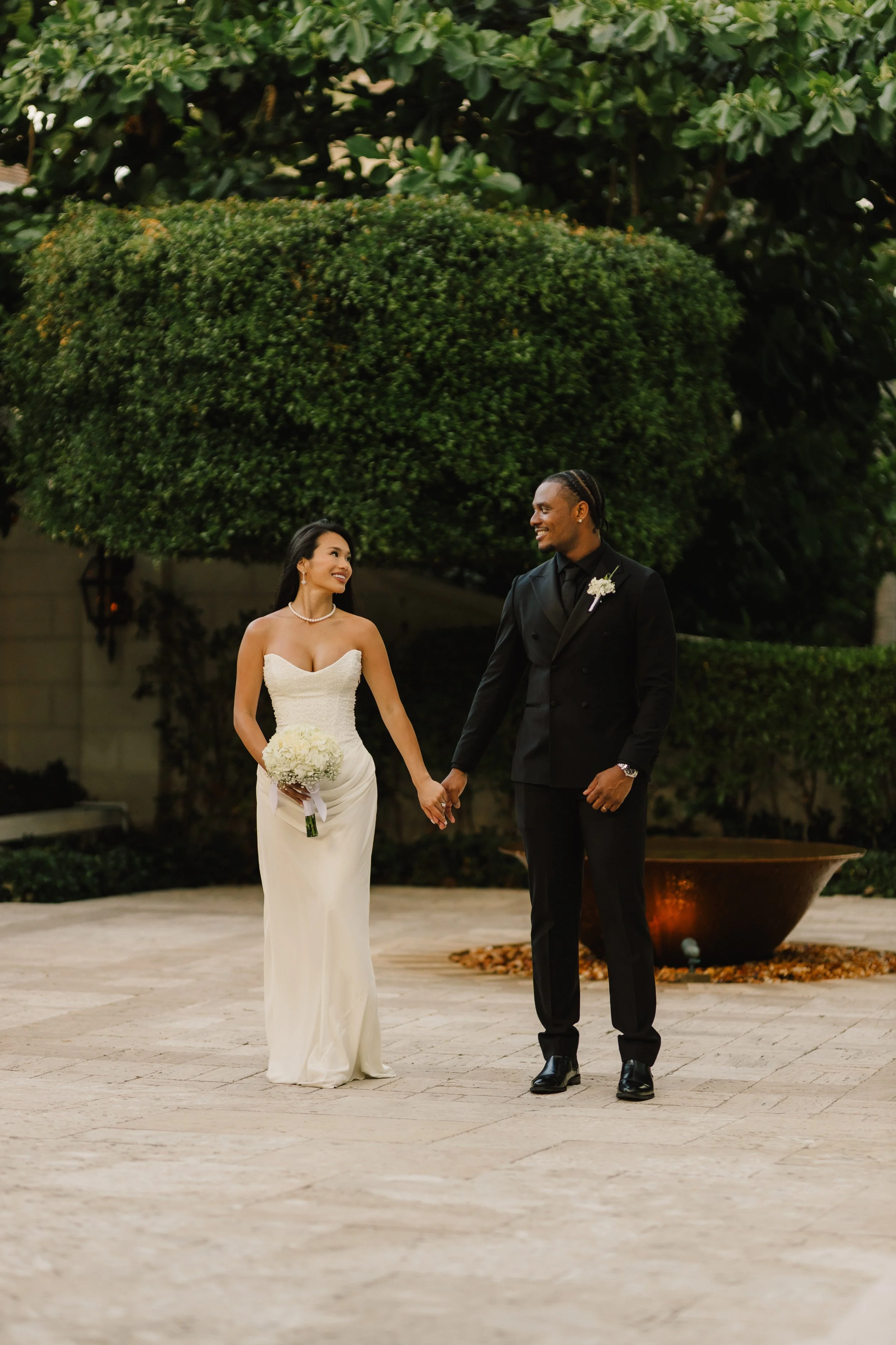 A bride and groom holding hands and looking at each other, smiling, outdoors during wedding. The bride wears a strapless white dress and holds a bouquet. The groom wears a black suit with a boutonniere. Large green trees are in the background.