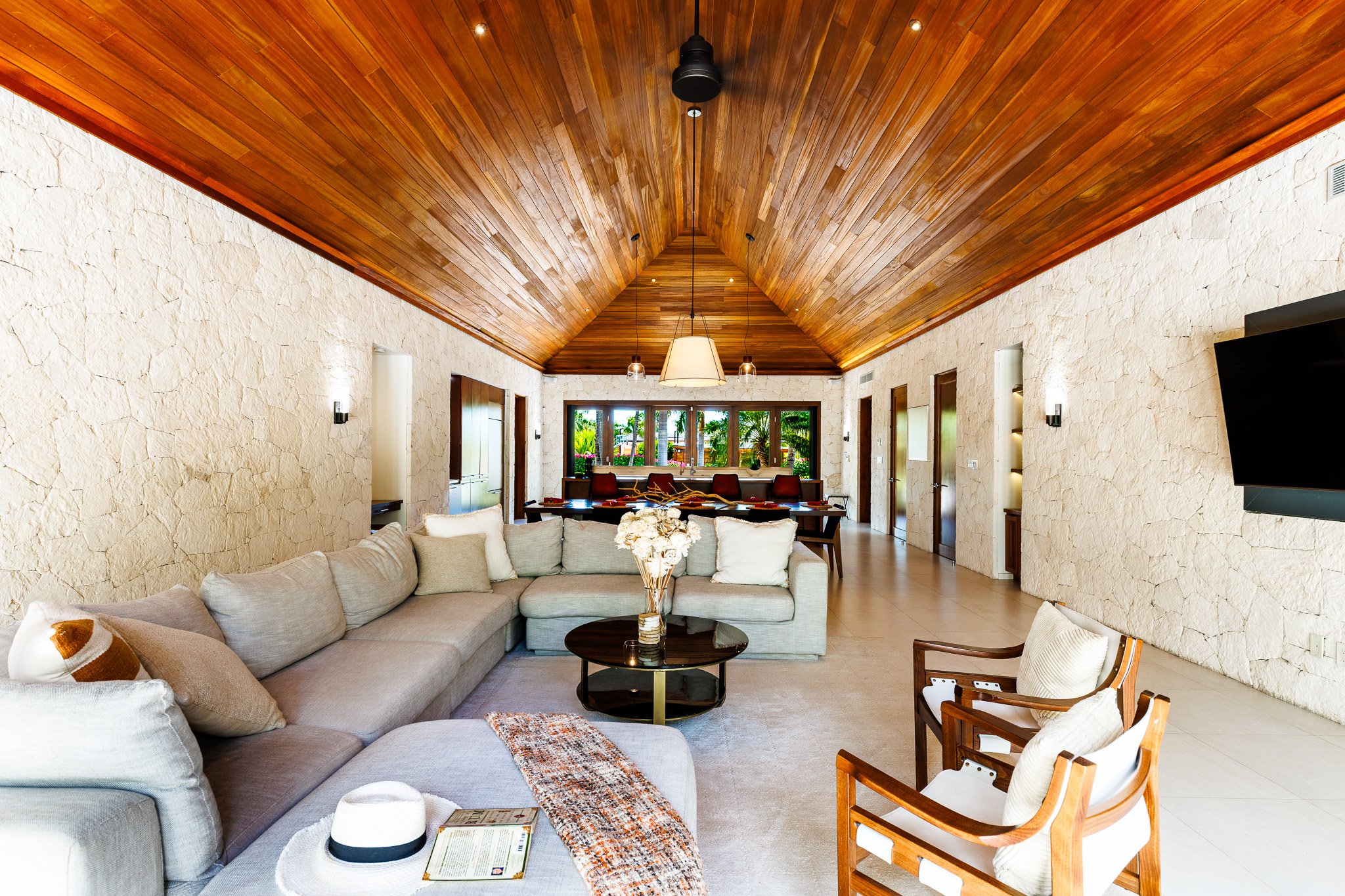 Living room with beige sectional sofa, a round coffee table with a vase of flowers, a wooden armchair, and a stone wall. The ceiling is made of wood with a central hanging lamp and spotlights. Large windows in the background showing greenery outside.